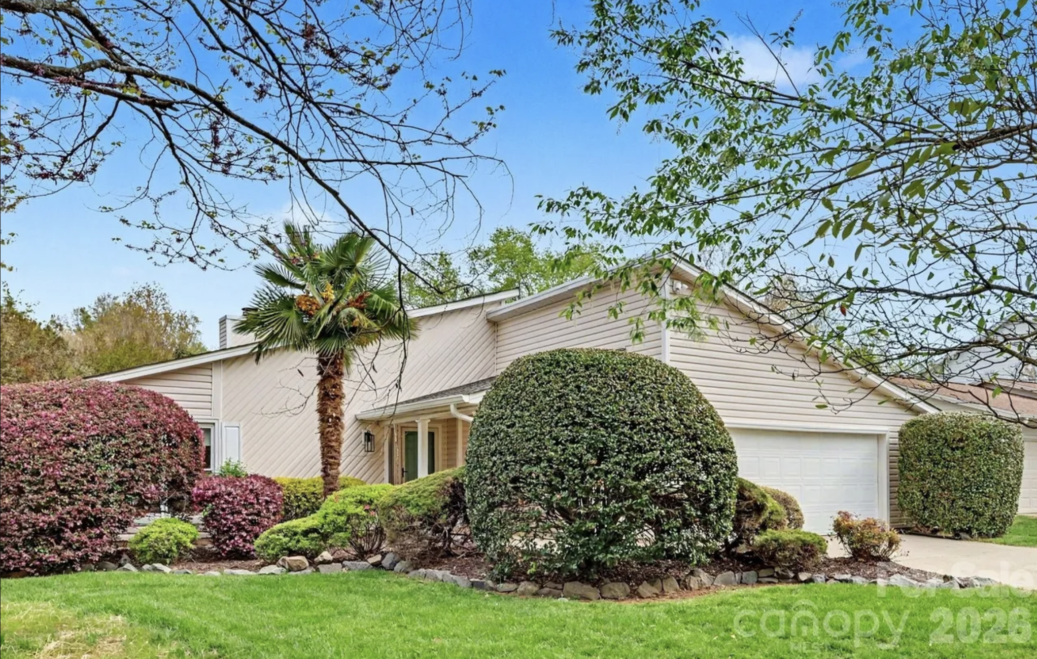 A single-story beige suburban house with a two-car garage, a neatly trimmed round shrub in the front yard, a tall palm tree, flowering bushes, and a green lawn under a clear blue sky.
