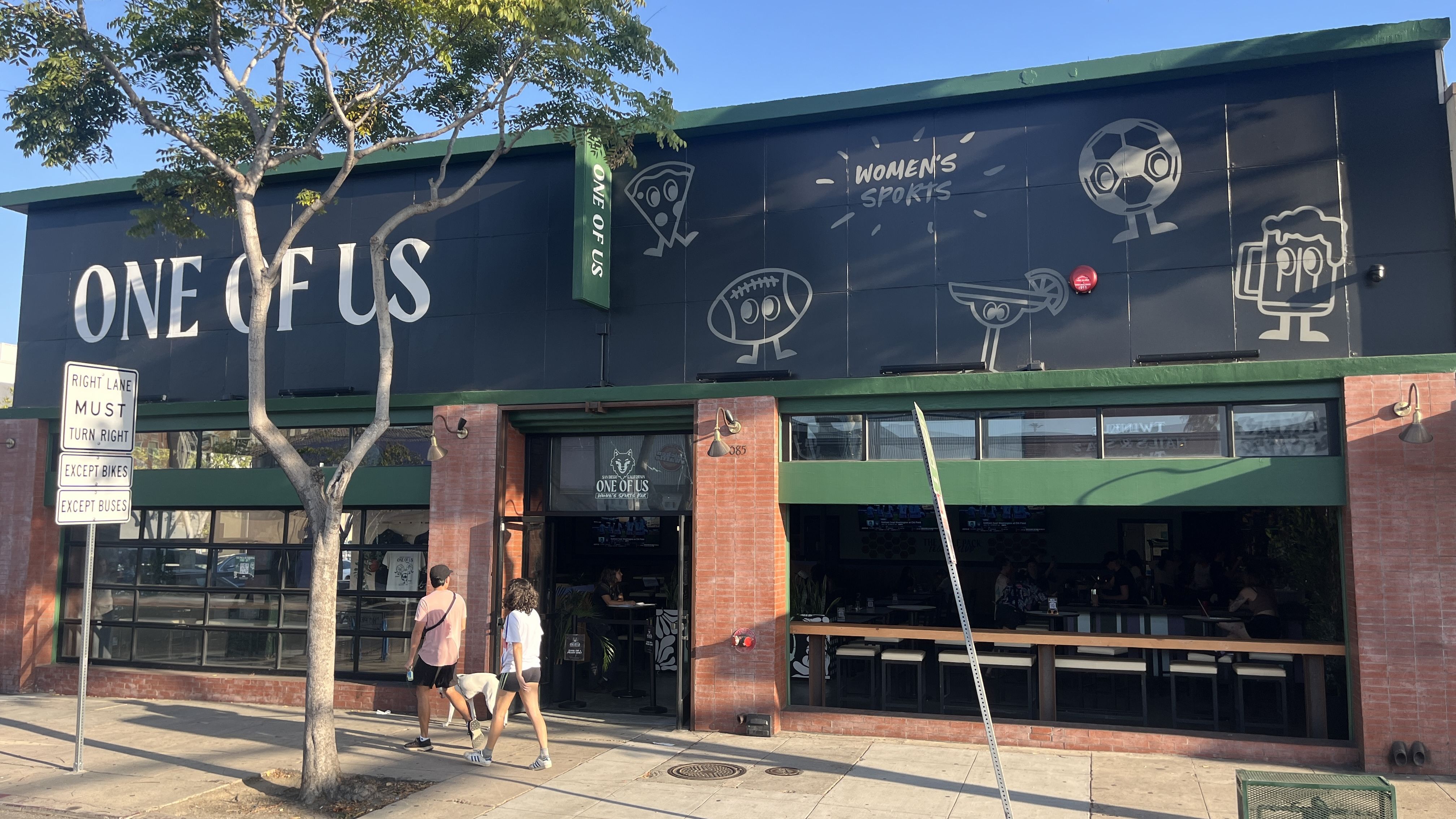 Exterior view of a sports bar with a dark wall reading "ONE OF US" in white letters. Line art shows pizza, football, beer mug, and "WOMEN'S SPORTS." Tree, pedestrians, brick facade, and green trim.