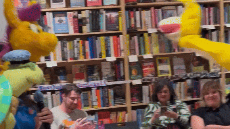 People at a book signing or reading event in a bookstore, with colorful costumed characters including a turtle and a yellow creature interacting with attendees.