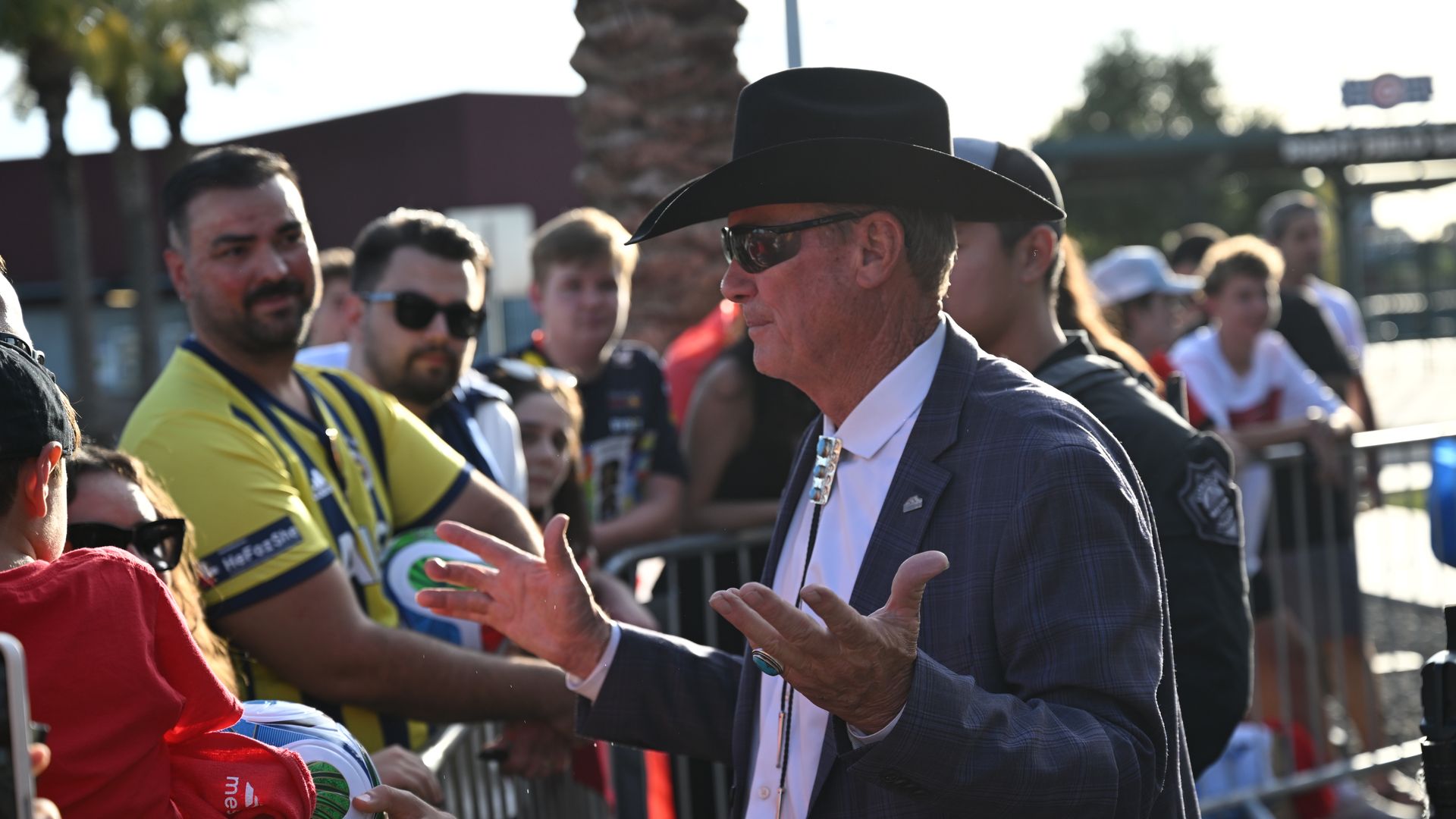 Man in a black cowboy hat and sunglasses, in a blue plaid suit, speaks to a crowd behind barriers as fans in yellow jerseys reach out on a sunny day.