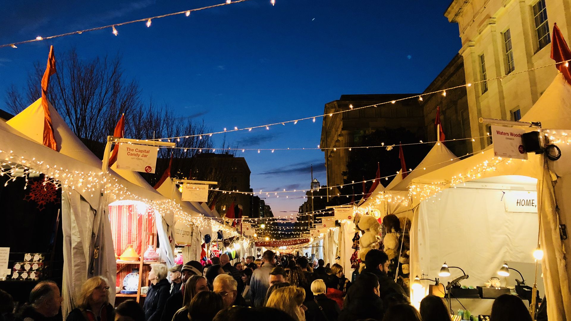 Evening market street lined with white tents and string lights, a crowd of people strolling between stalls. A grand lighted building on the right, dark blue sky, leafless trees in distance.