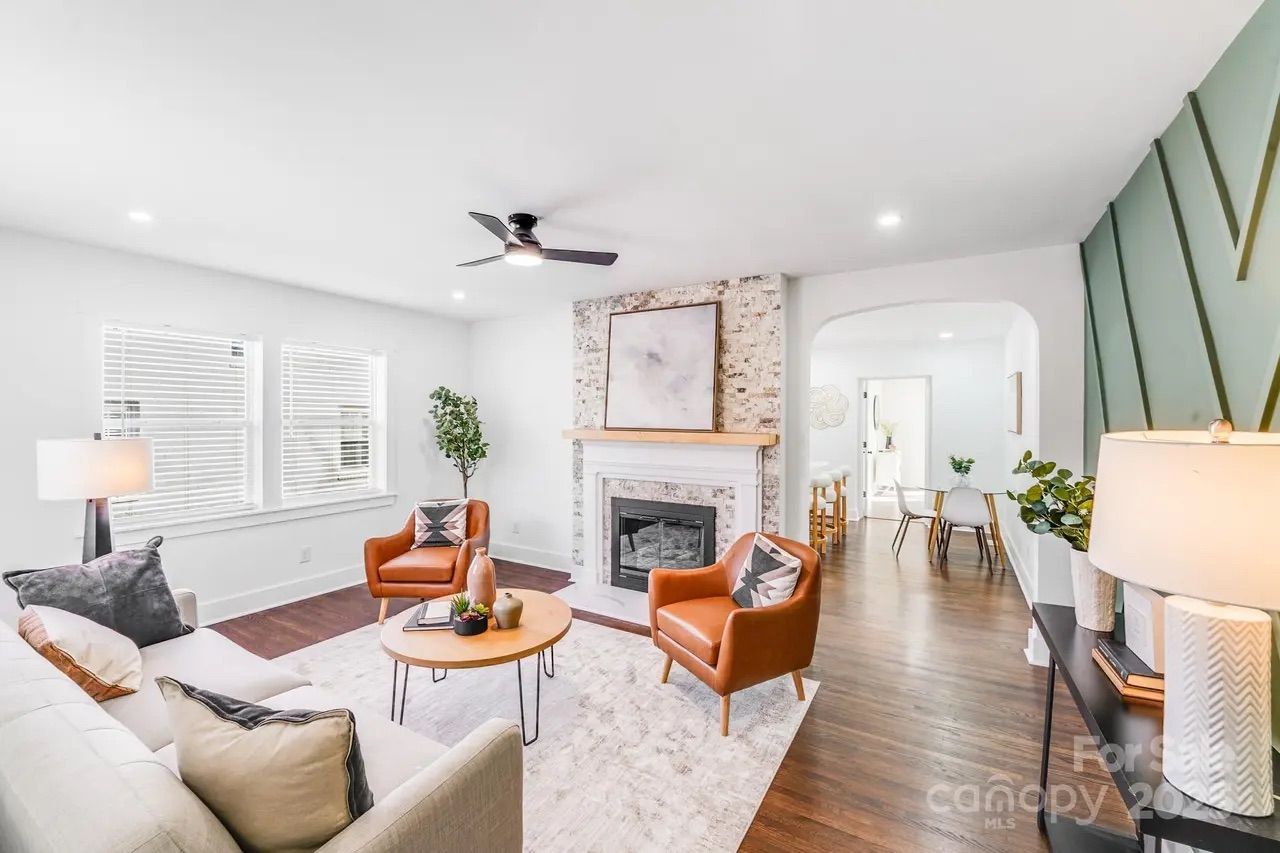 Bright living room with white walls, a brick fireplace, two brown leather armchairs, a beige sofa, wooden coffee table, hardwood floors, and a ceiling fan. Adjacent dining area visible.
