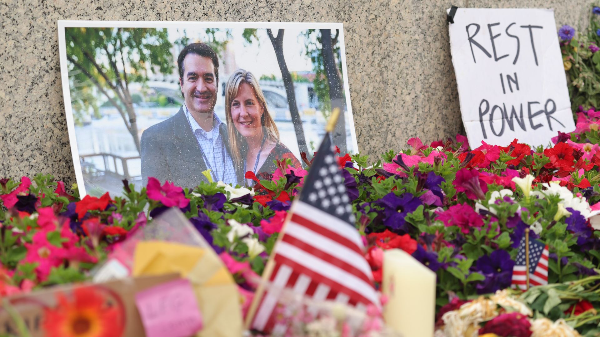 Memorial scene with a framed photo of Mark and Melissa Hortman leaning against a granite wall, surrounded by colorful flowers and small American flags; a white sign reads REST IN POWER.