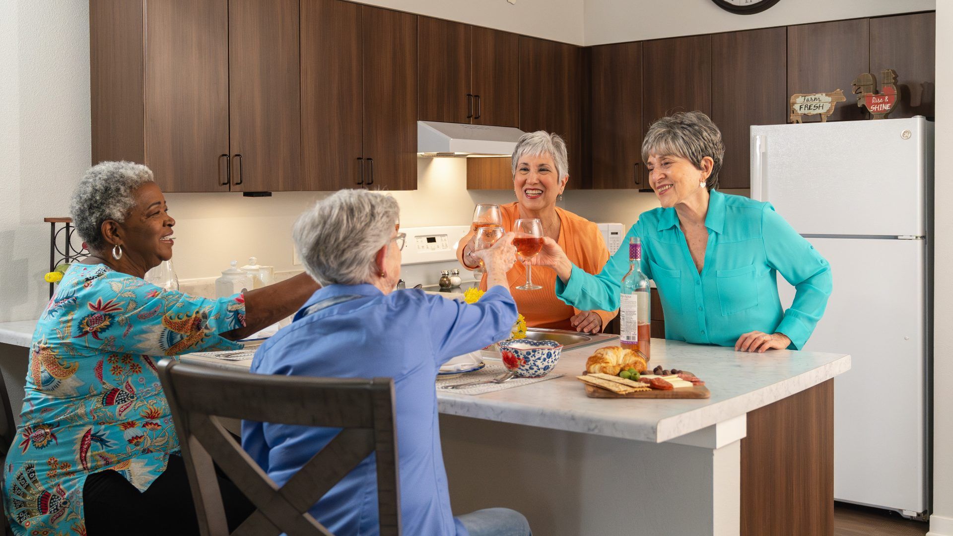 A group of four older women gather around a kitchen island, raising glasses of rosé wine in a cheerful toast.