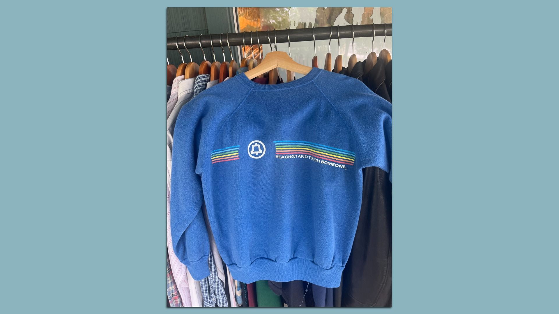Blue sweatshirt on a hanger in a clothing rack, with rainbow stripes and a white circular logo; the slogan "REACH OUT AND TOUCH SOMEONE" is printed on the back.