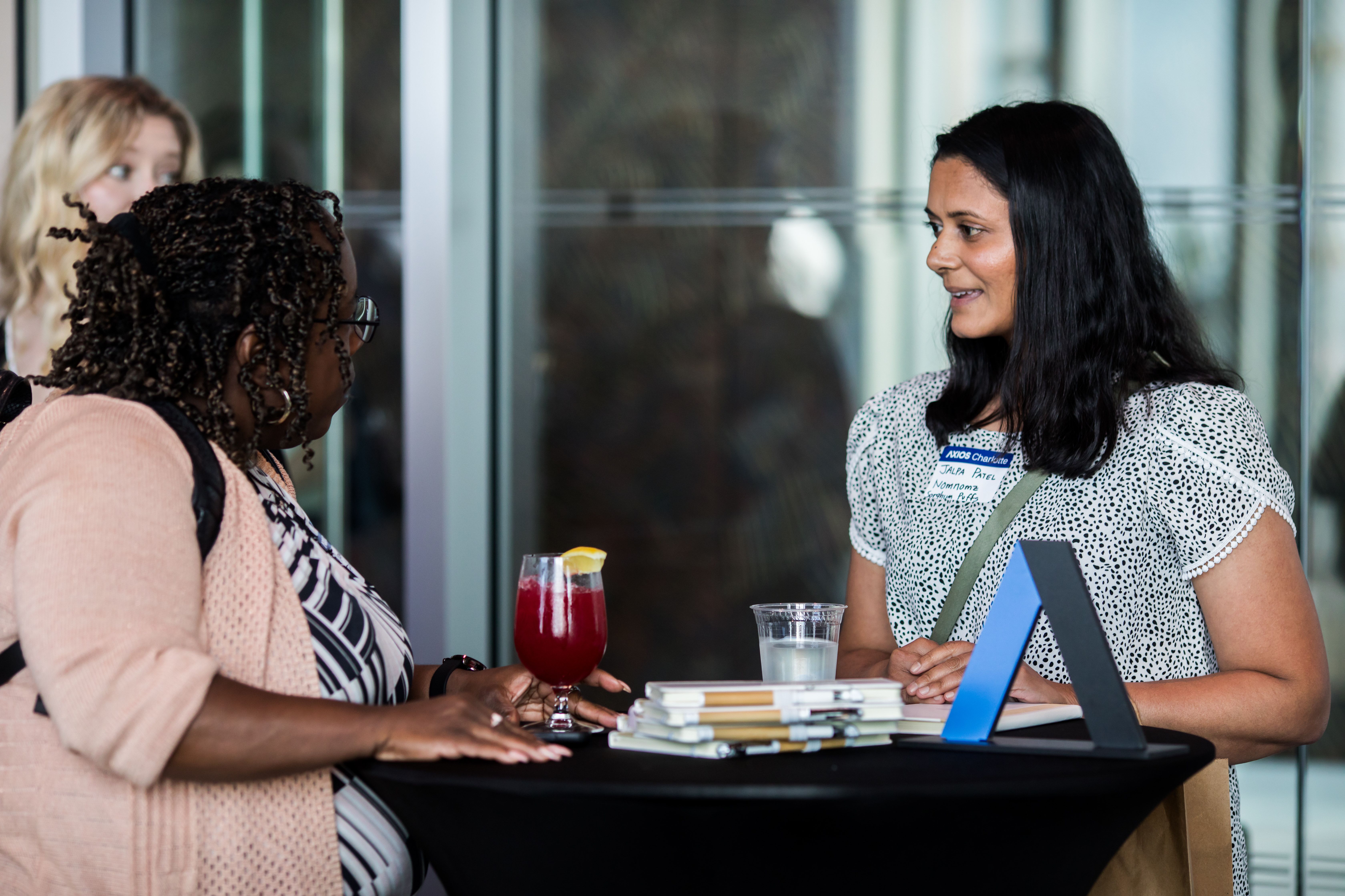 Two women chat at a round black table: one with light hair and a pink cardigan, the other with long dark hair and a name badge. On the table are a red cocktail, a clear drink, and a stack of magazines.