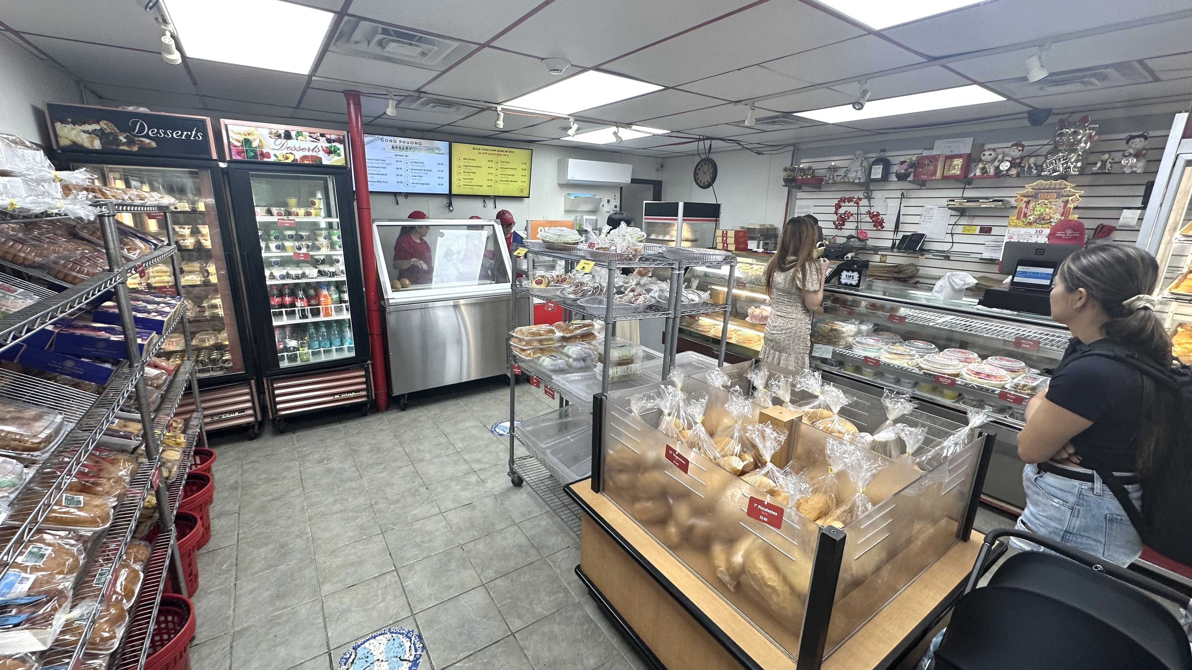 Interior of a bakery with shelves and racks filled with packaged bread and desserts, a refrigerated drink case, menu boards, and two customers at the counter with staff behind it.