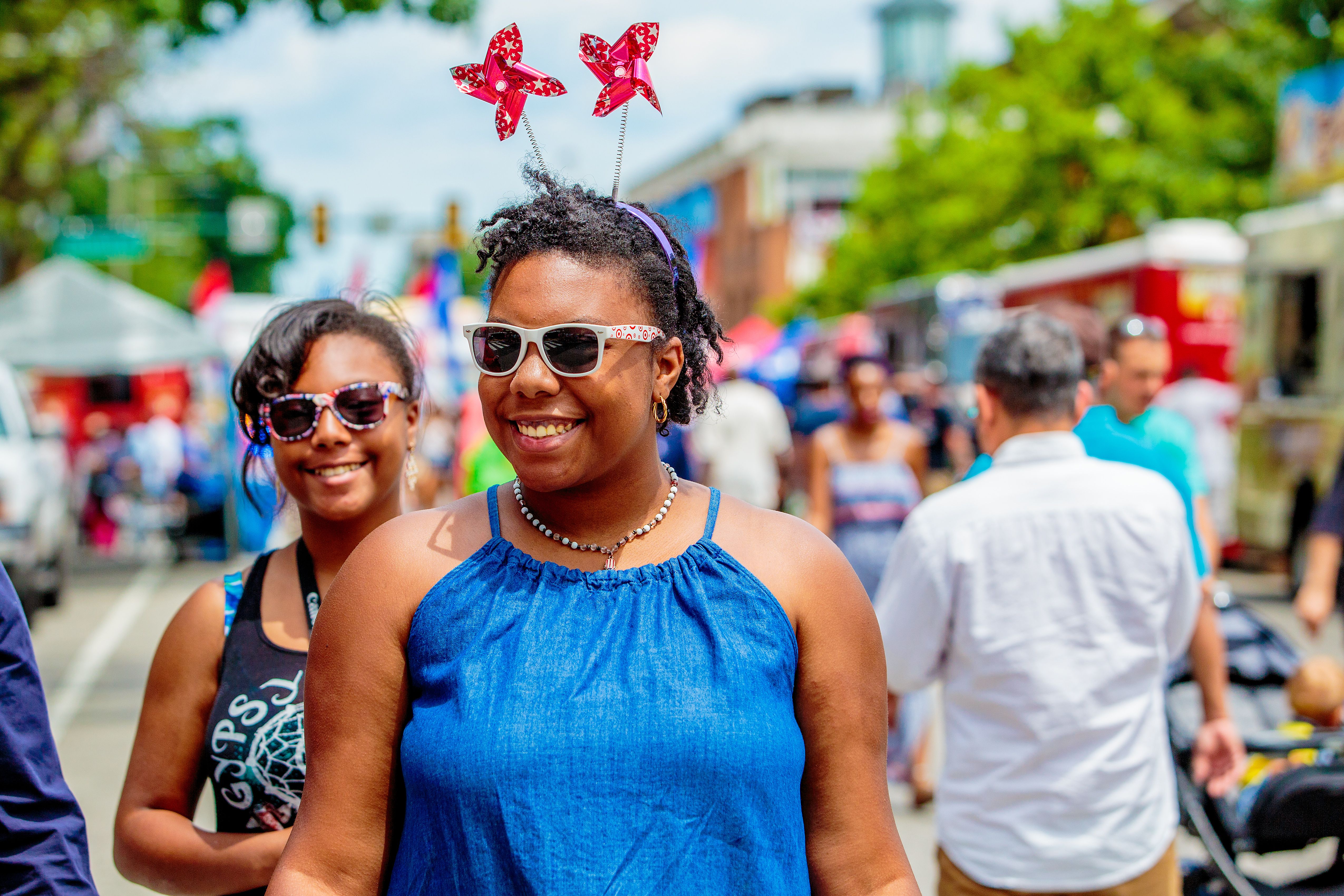 A woman in a sleeveless shirt wearing sunglasses, a necklace and a headband with pinwheels on the top amid a festival. 