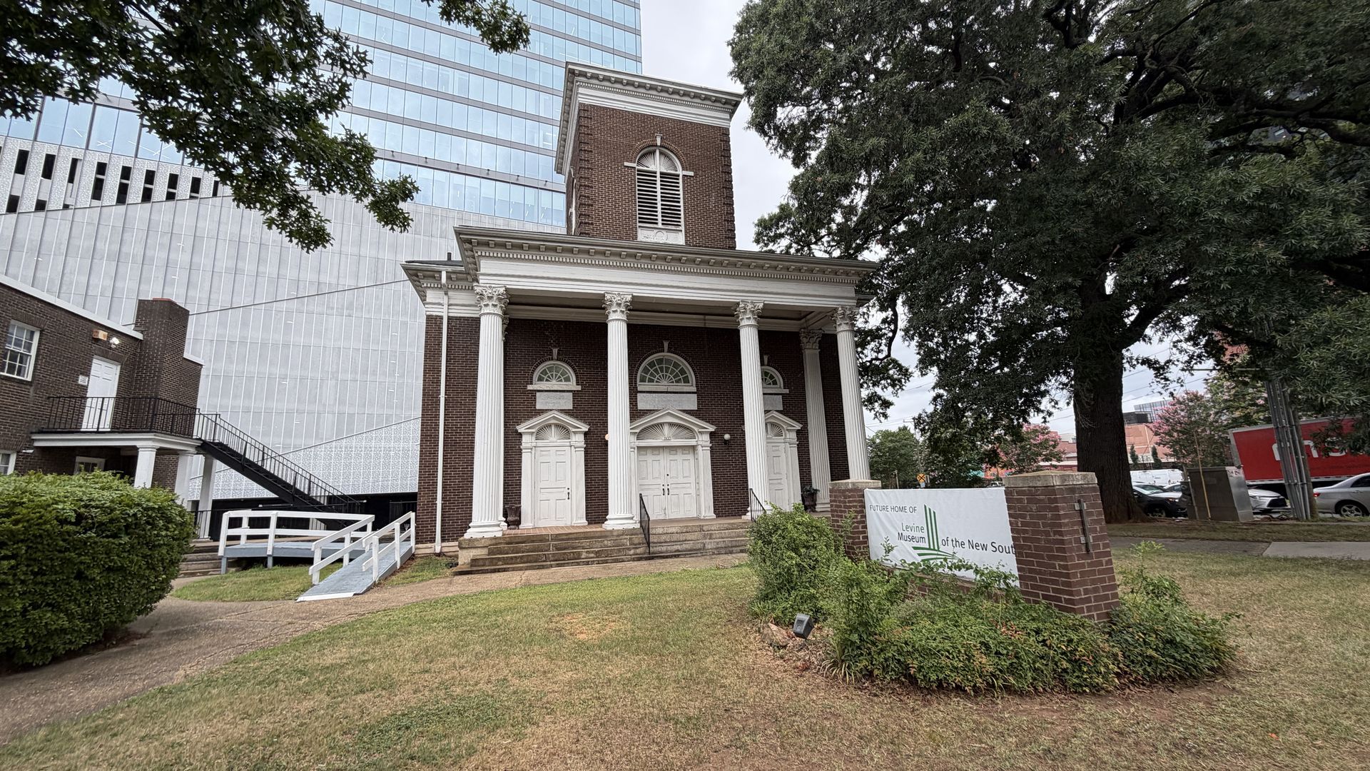 Historic brick building with white Doric columns and a pedimented porch stands beside a modern glass building; a large tree on the right and a sign for the Levine Museum of the New South.
