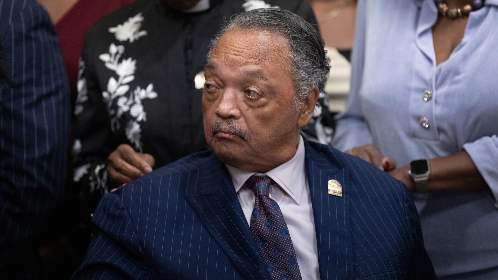 A sitting civil rights leader Rev. Jesse Jackson, wearing a navy pinstriped suit, lilac shirt and brown-purple tie with blue dots on it looks to his right across a crowded room.
