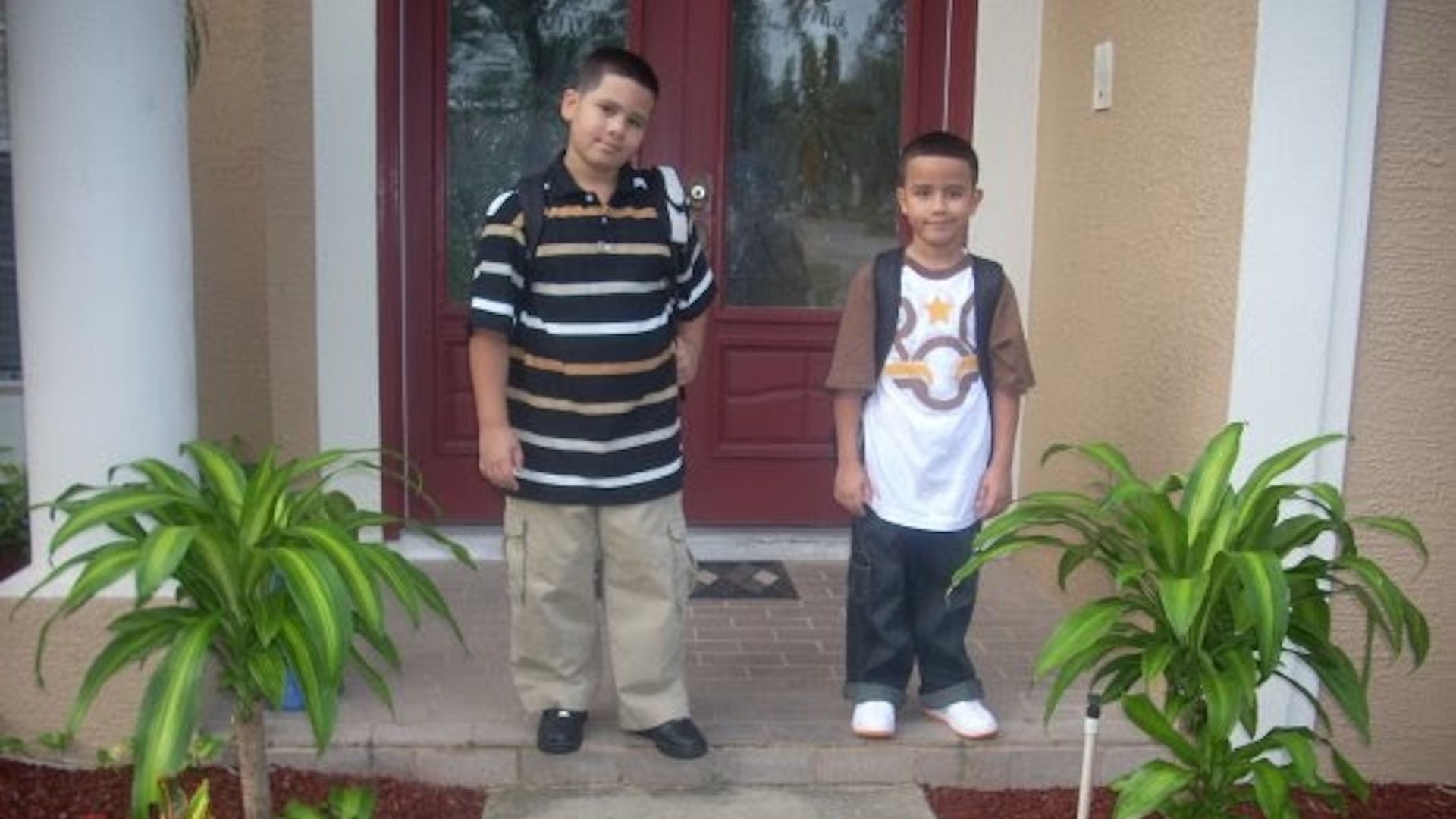 Two young boys with backpacks stand on a front porch in front of a beige house with a red door; one wears a black striped polo and tan pants, the other a white and brown T‑shirt with jeans, flanked by green plants.