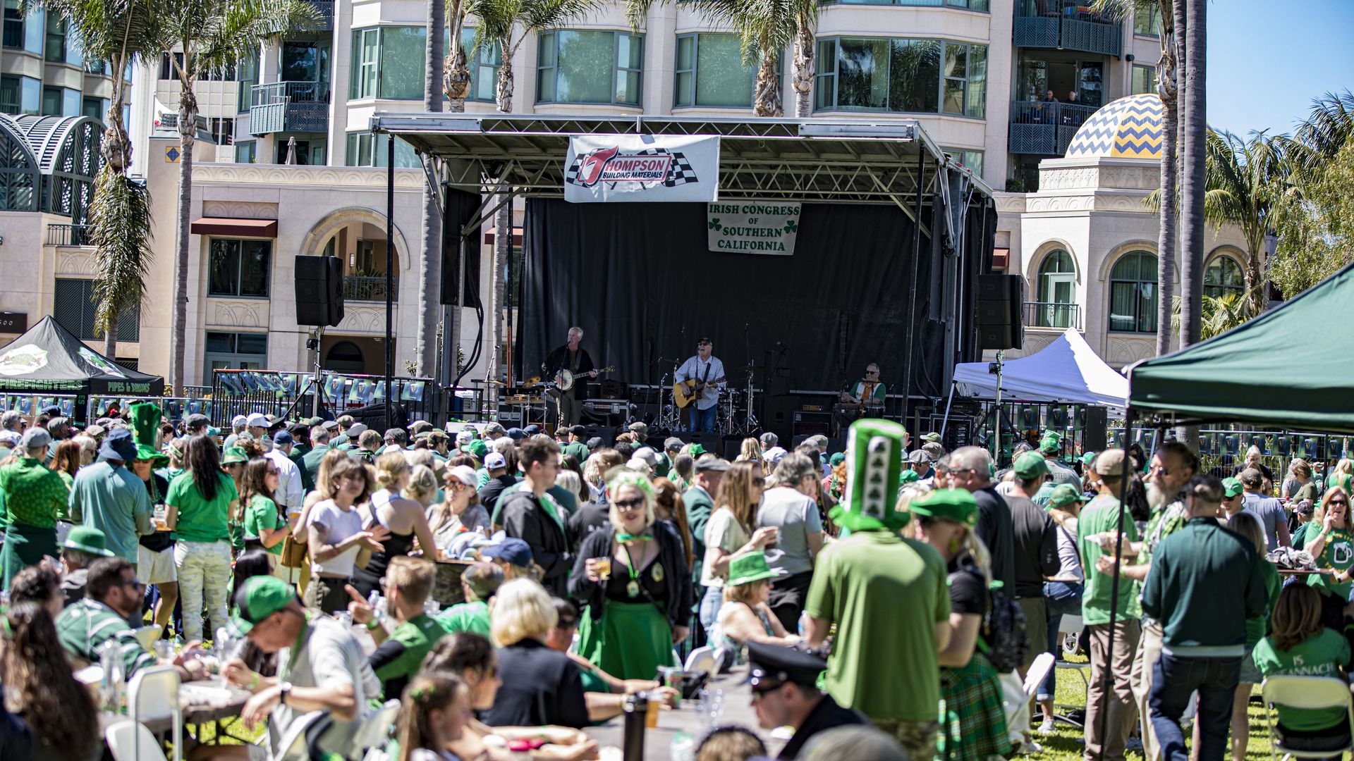 A crowd of people dressed in festive green Saint Patrick's Day attire eat and gather around a live music stage at Balboa Park. 