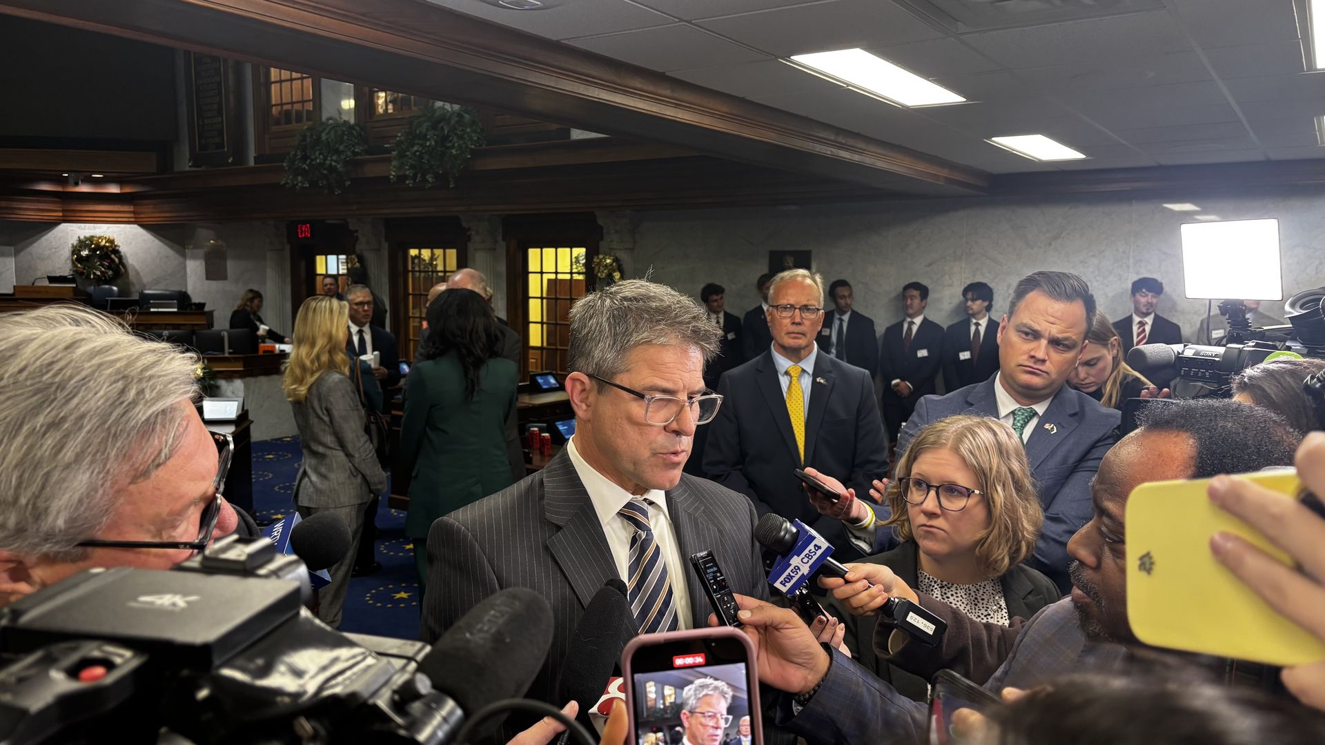 Man in striped suit and glasses speaking to reporters with microphones and cameras in an indoor formal room with blue carpet and wooden trim.