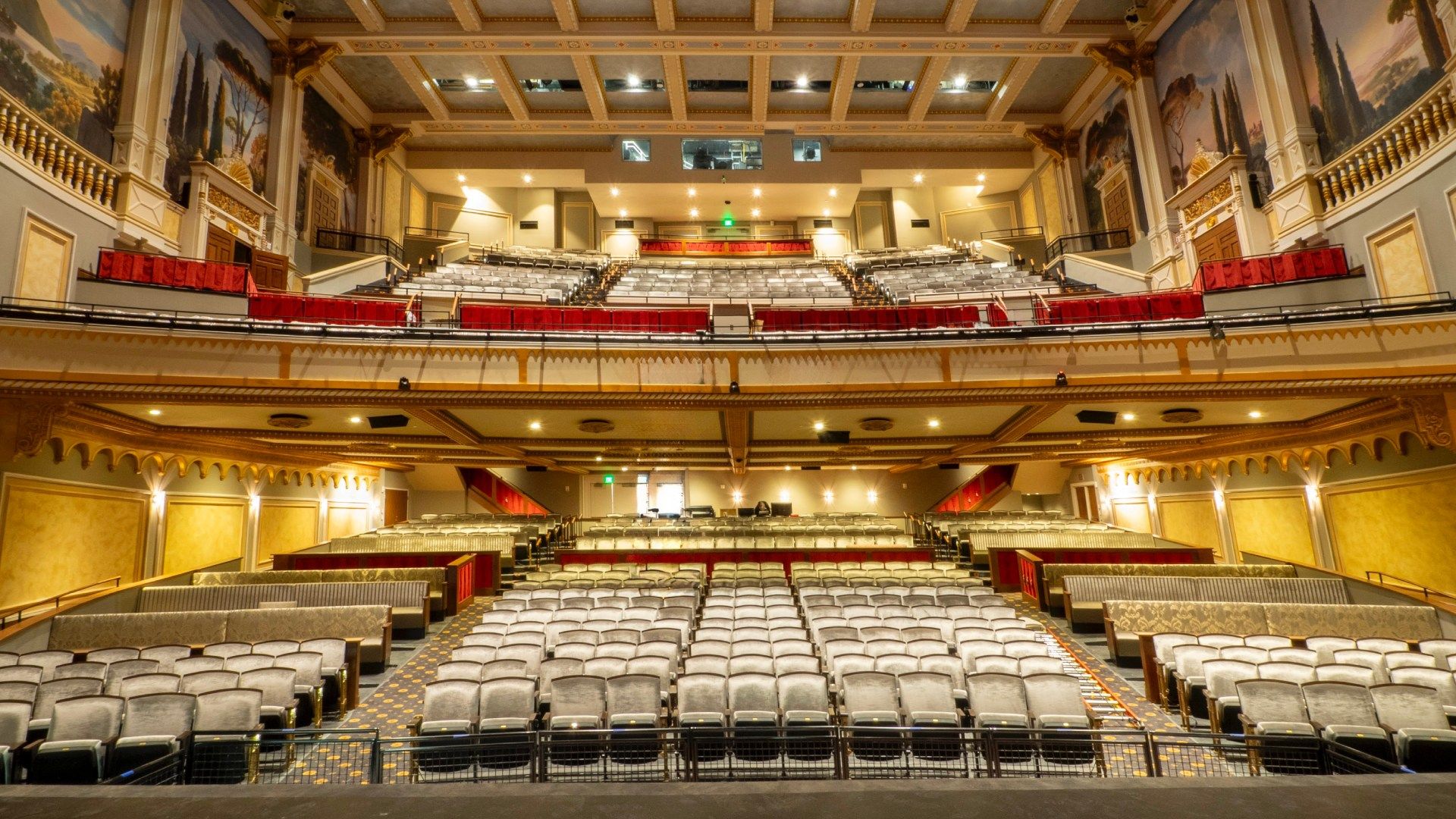 Interior view of a grand theater with tiered gray seating, ornate beige and gold walls, red balconies and large murals on the upper walls under a decorative ceiling.