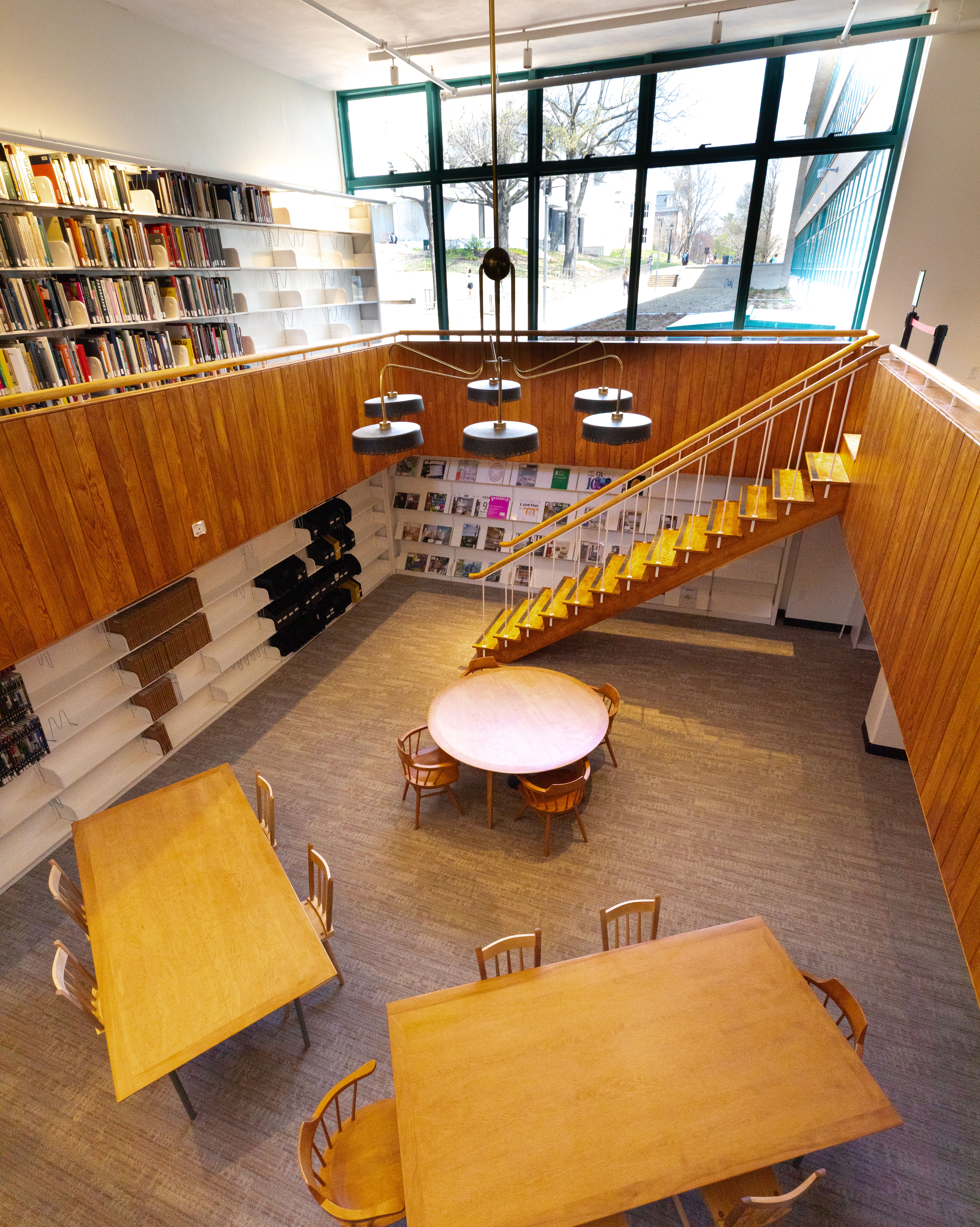 A bright library interior with wooden tables, bookshelves, a staircase, and large windows overlooking an indoor courtyard.