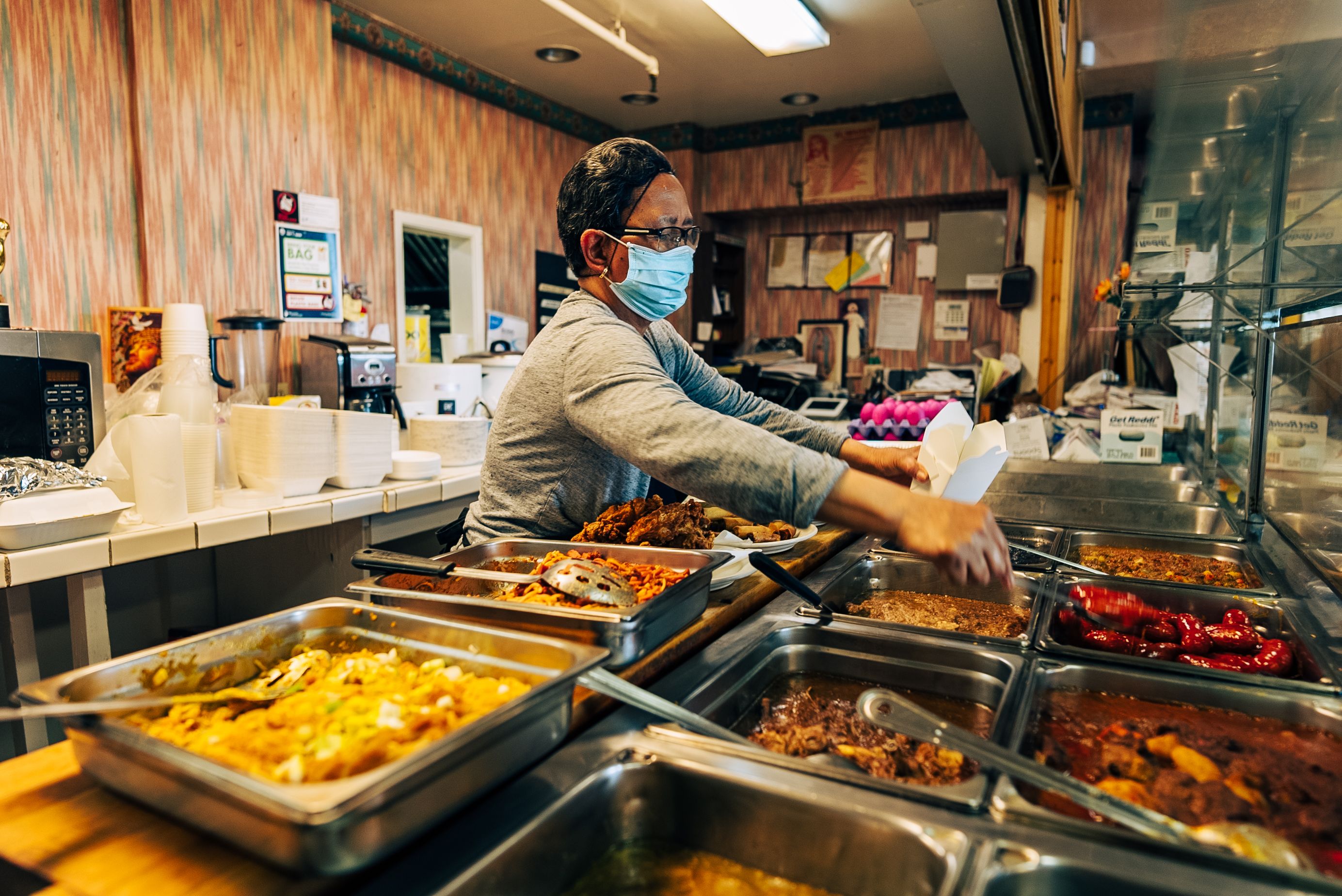 Person wearing glasses and a blue face mask serving food from a buffet with various dishes in metal trays inside a restaurant kitchen with wood panel walls.