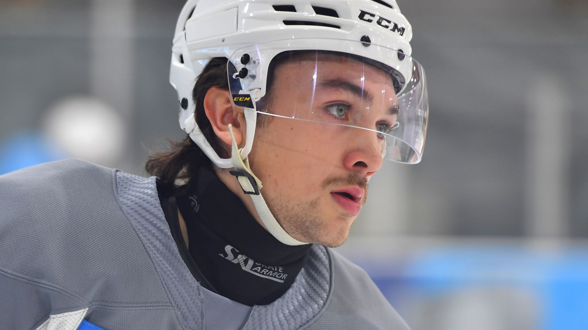 A hockey player with light skin and a short moustache in a helmet wears a neck guard — a black piece of cut-resistant fabric that fits closely to his neck.