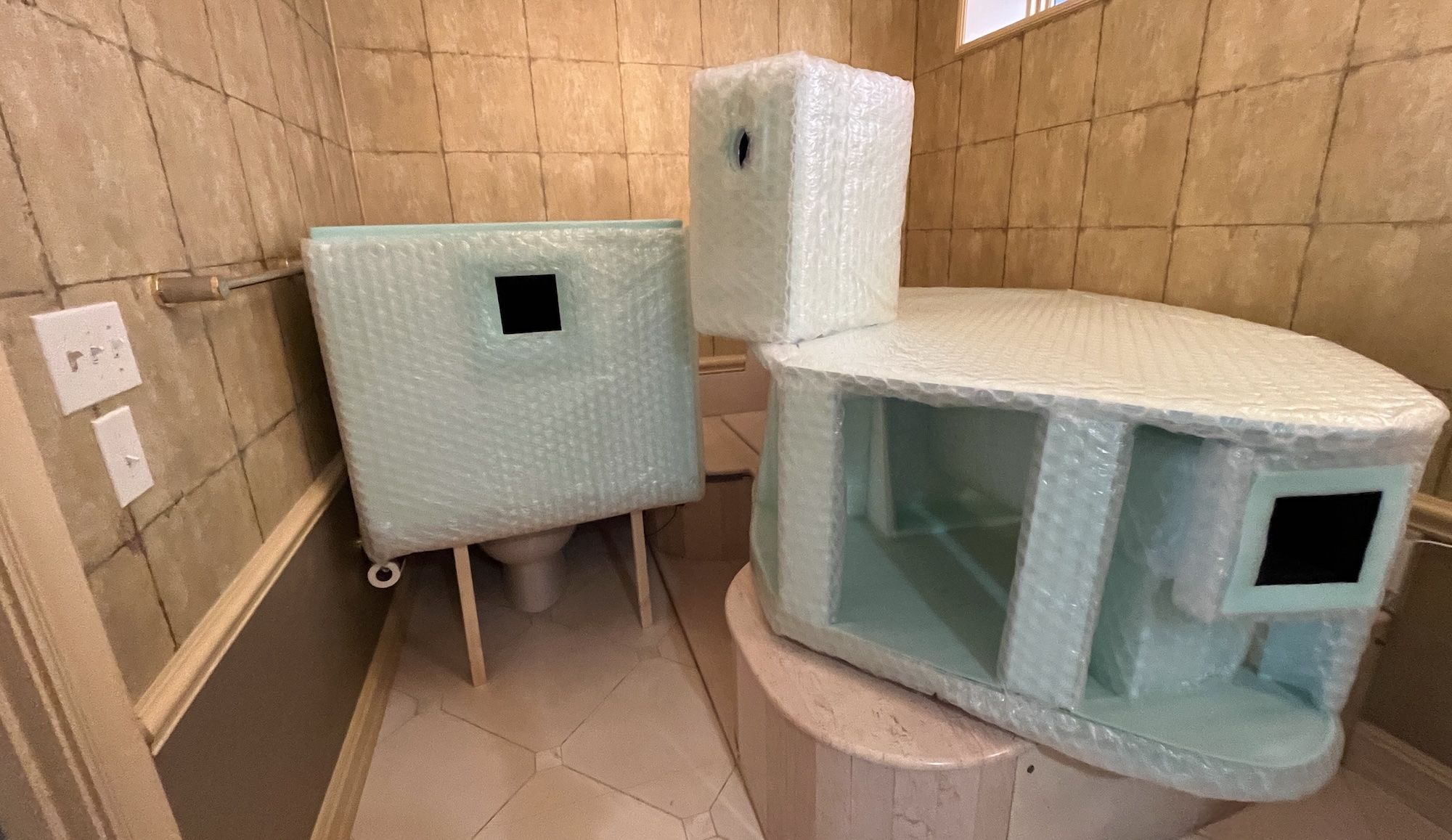 Bathroom with tan tile walls. Bubble-wrapped pale turquoise foam props include a curved, tub-like unit with an open front and a rectangular box with a square window; a tall foam block stands nearby.