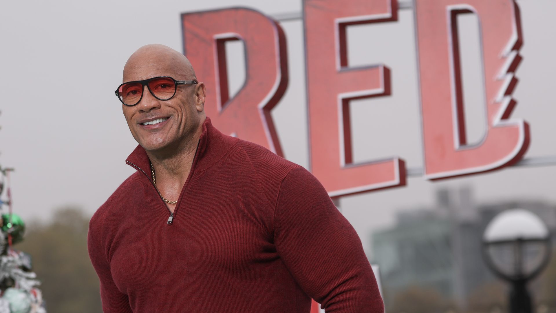 Dwayne "The Rock" Johnson in a red quarter zip with the collar up in red see through glasses smiling at the camera. The word "RED" is behind him