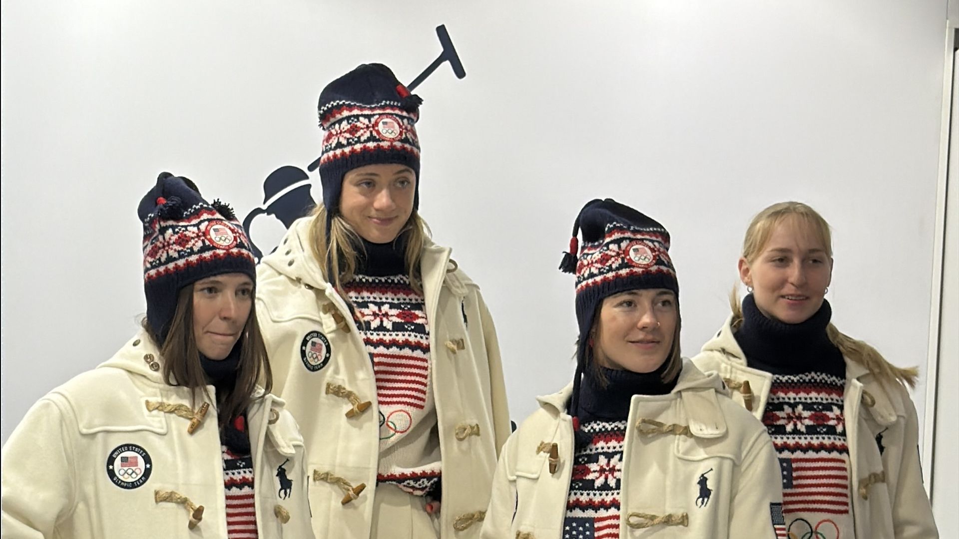 Four women dressed in matching white coats, pants, and knit hats with red, white, and blue patterns, posing indoors against a white background with blue stars, wearing brown boots.