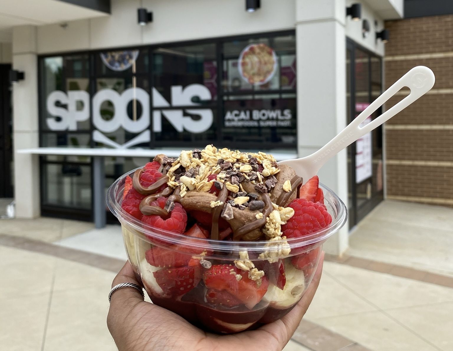 Hand holding a clear bowl of acai topped with granola, raspberries, strawberries, bananas, almonds, and chocolate drizzle outside a shop with "SPOONS Acai Bowls" signage.