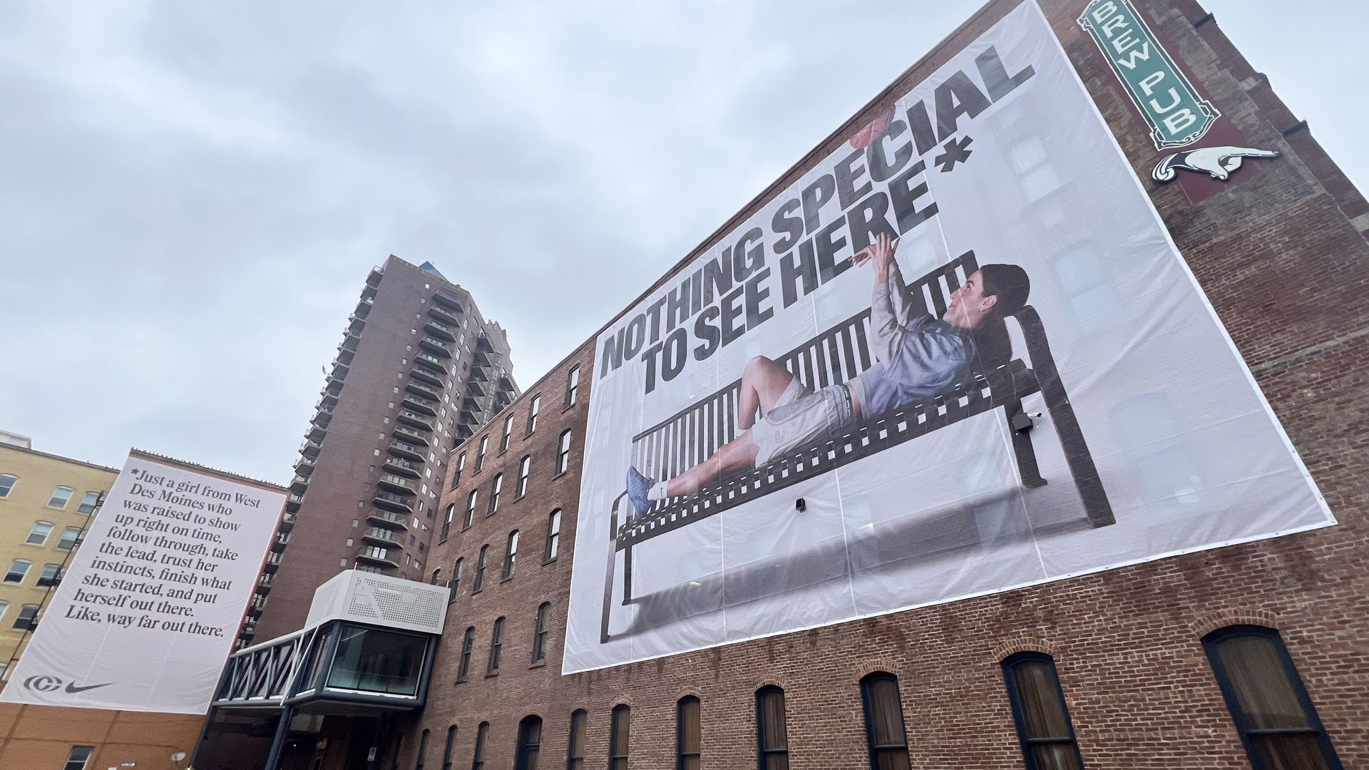 Large outdoor banner on a brick building shows a person reaching up on a film-strip graphic with the words "NOTHING SPECIAL TO SEE HERE." A green "BREW PUB" sign hangs at the corner; cloudy sky overhead.