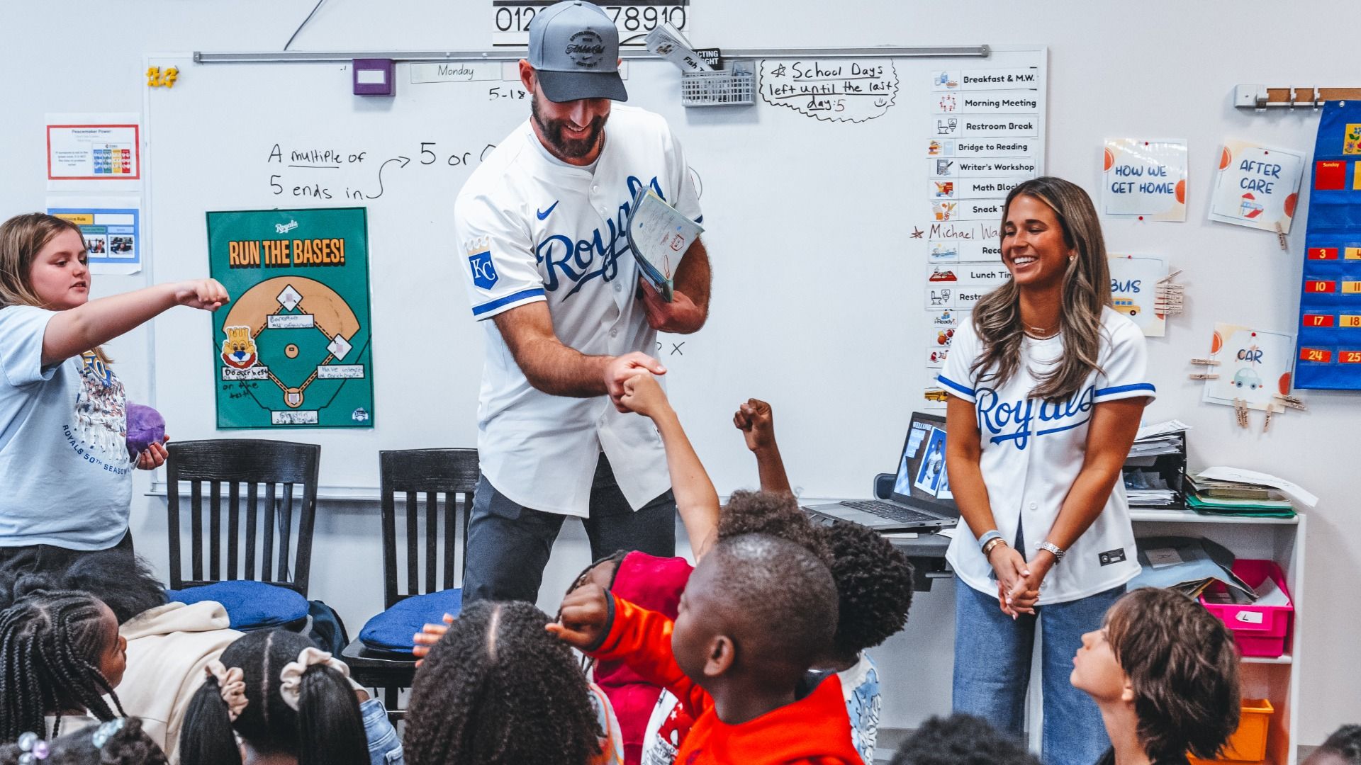 Classroom scene with a male Royals baseball player and a smiling female coach giving high-fives to kids. A whiteboard shows math notes and a 'RUN THE BASES!' poster.