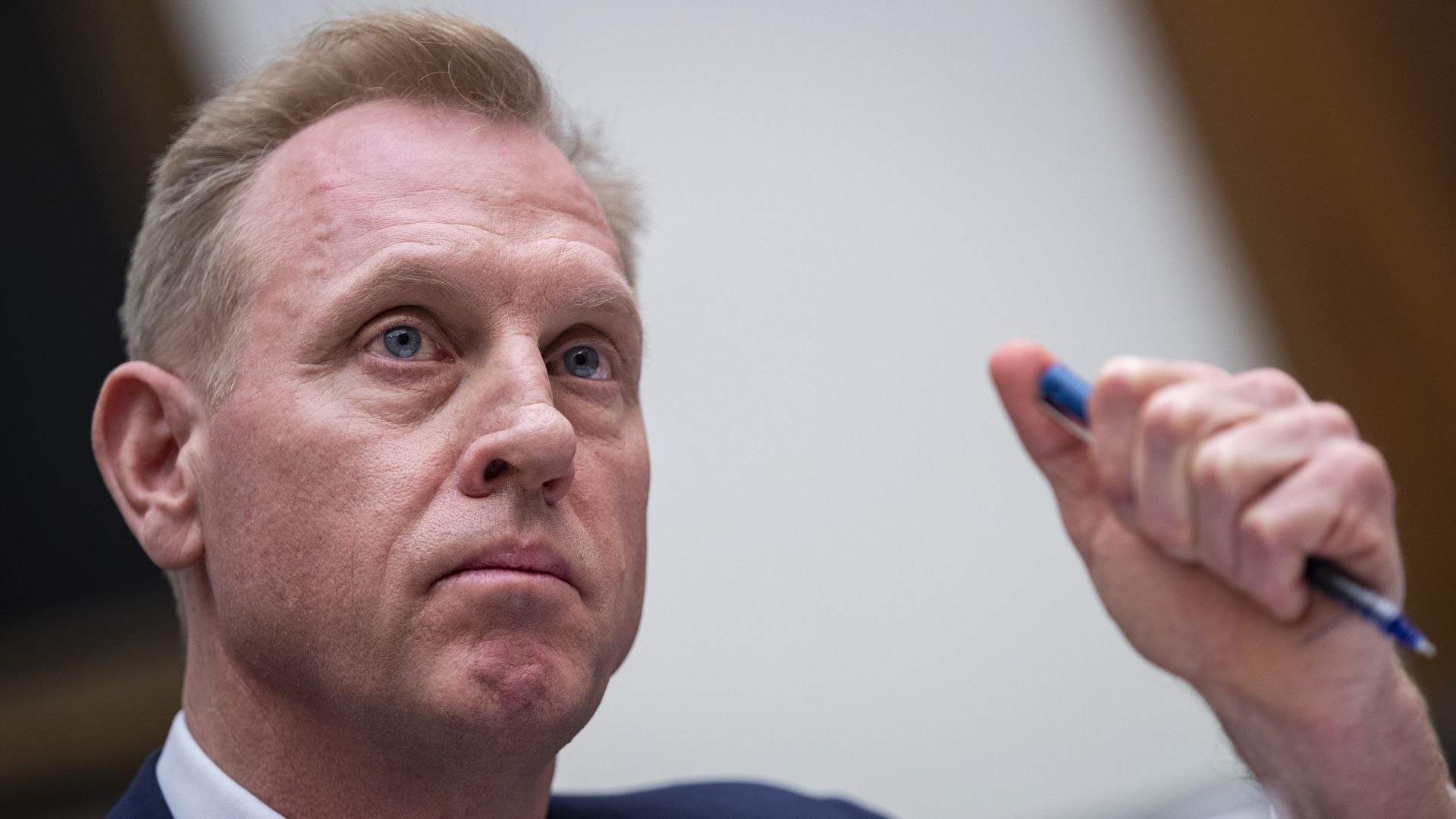 In this image, Patrick Shanahan sits and frowns while holding a pen in his fist.