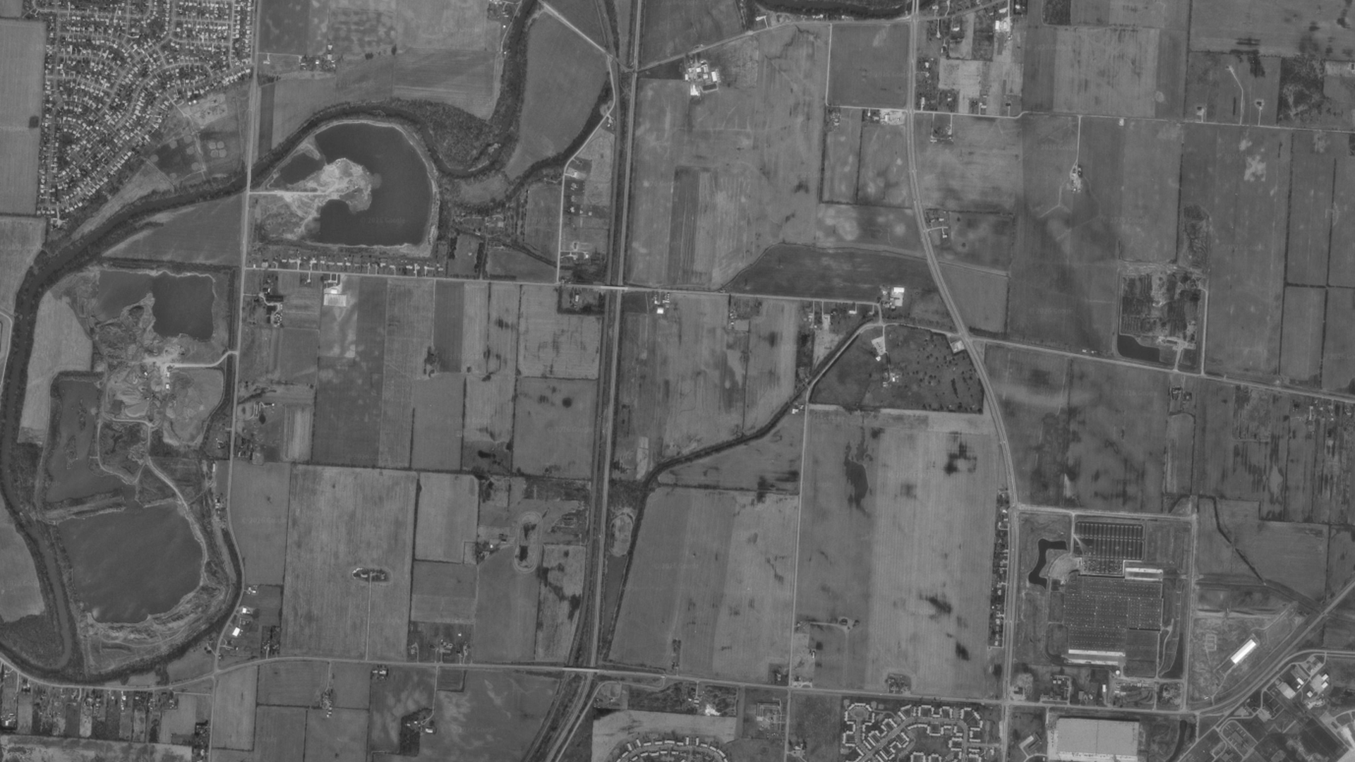Grayscale satellite view of a rural landscape: a winding river on the left, several ponds, patchwork fields, and roads with scattered houses. A large rectangular facility sits bottom-right.