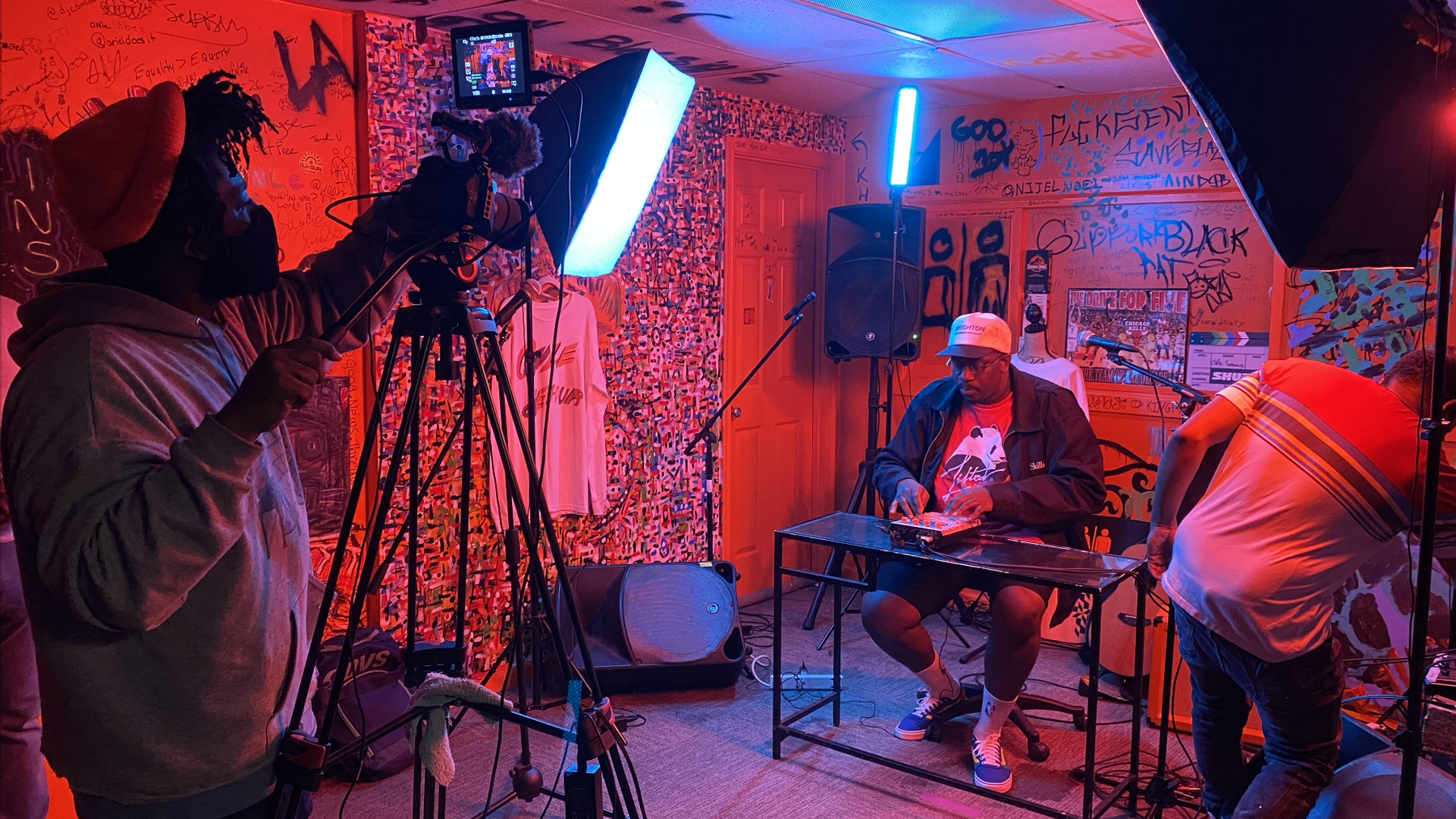 A man operates a camera as a musician plays an electronic sound board under red/orange lighting.