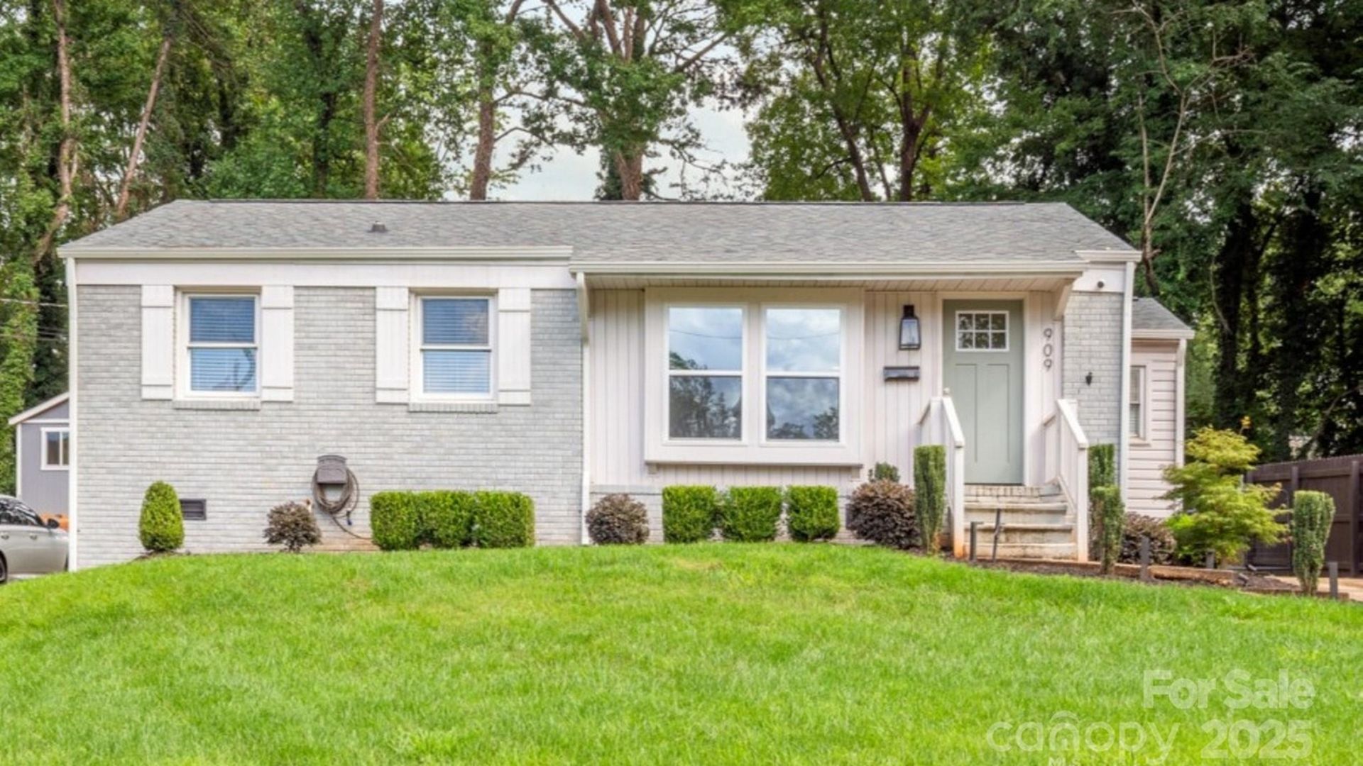 Single-story gray brick house with green front door, white-trimmed windows, small front porch, manicured bushes, green lawn, and tall trees in the background.