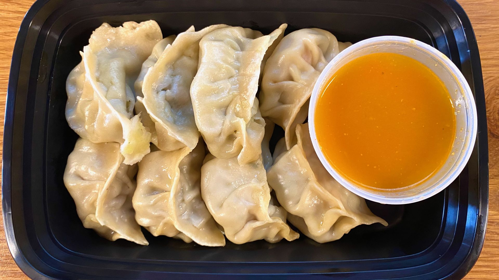 Eight dumplings in a black takeout container with a cup of orange momo sauce
