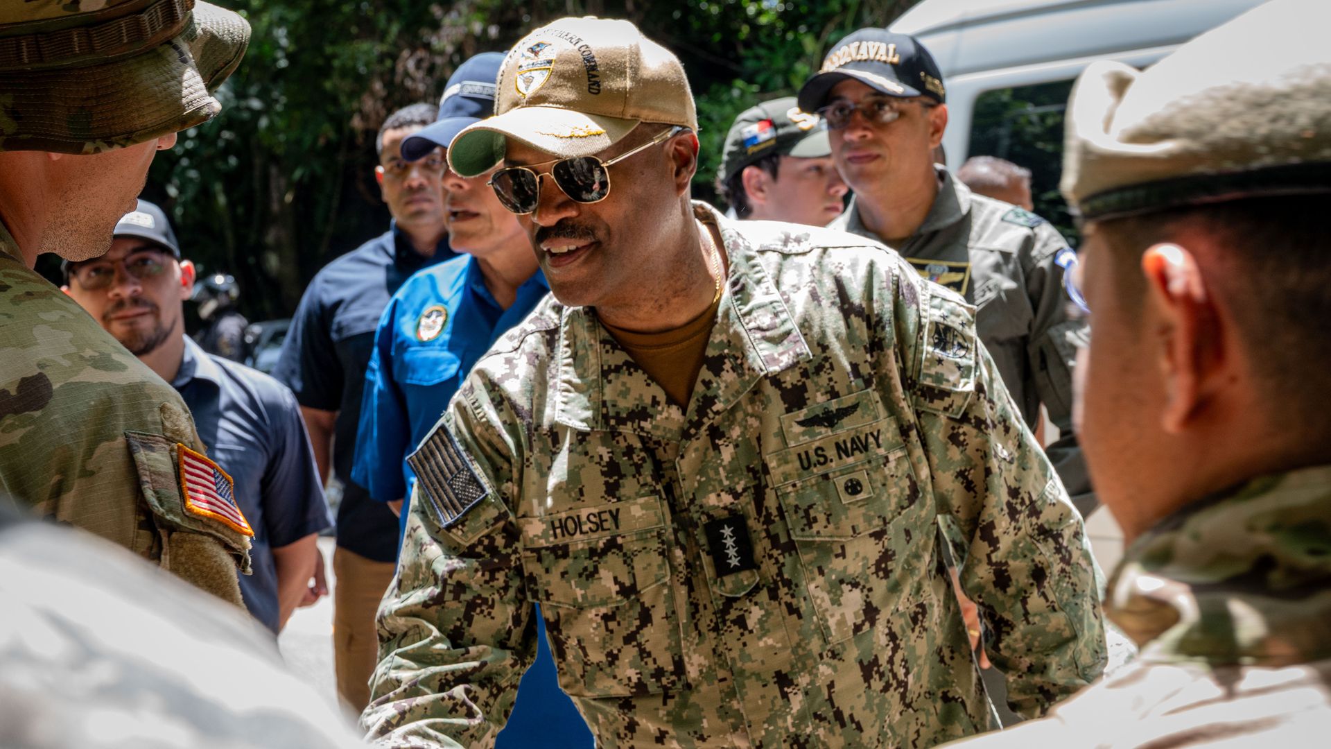 U.S. Navy officer in camouflage uniform and sunglasses shaking hands with another soldier outdoors, surrounded by military and civilian personnel.