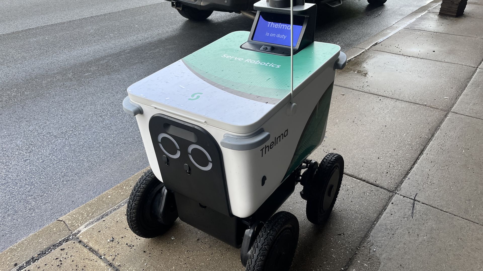 Autonomous delivery robot named Thelma on a wet city sidewalk, screen reads "Thelma is on duty." Green and white platform with four wheels; a black pickup is in the background.