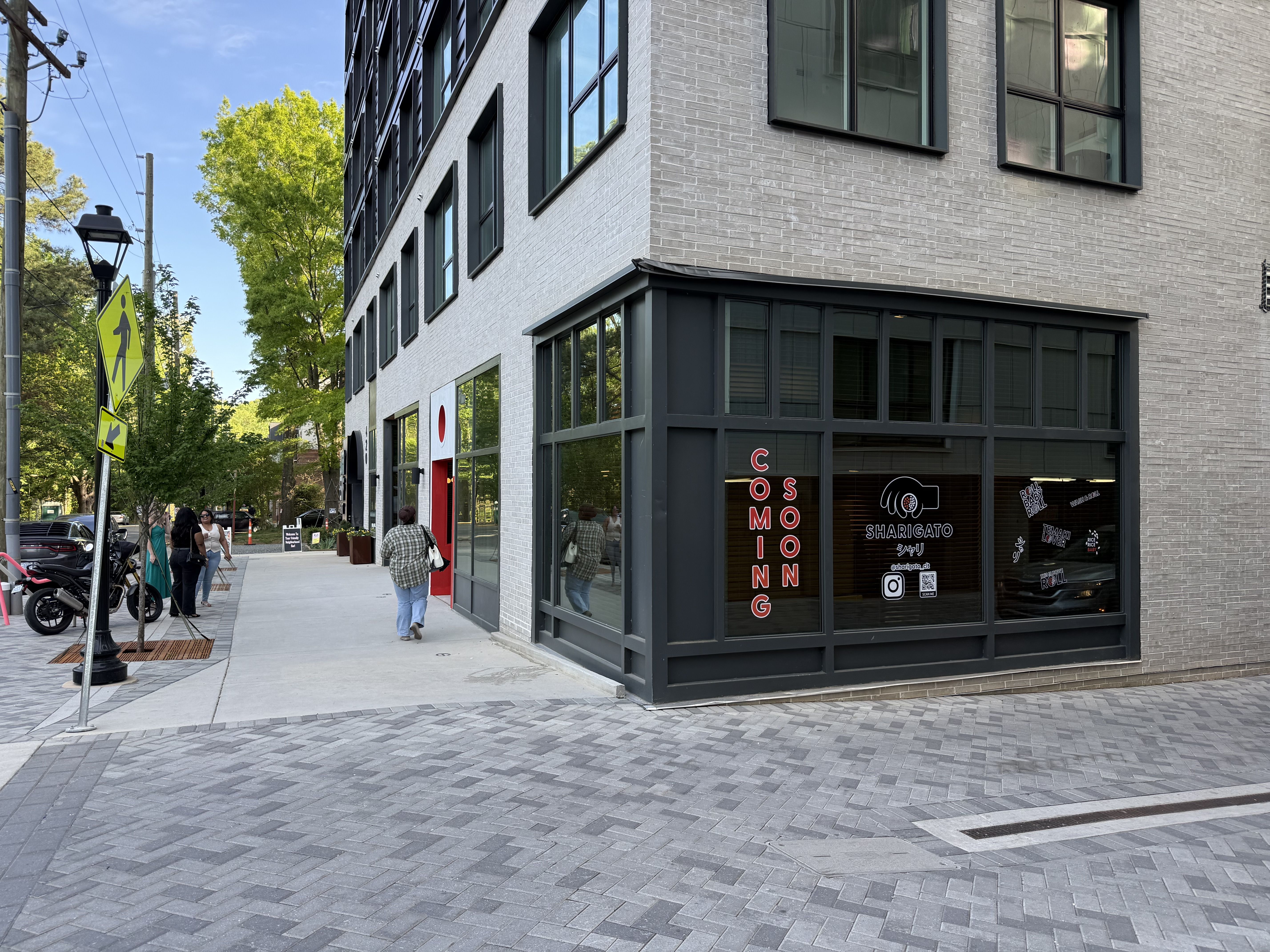 Corner view of a modern gray brick building with large black-framed windows; storefront shows a red vertical "COMING SOON" sign and logo; pedestrians stroll the sidewalk near a yellow crosswalk sign.