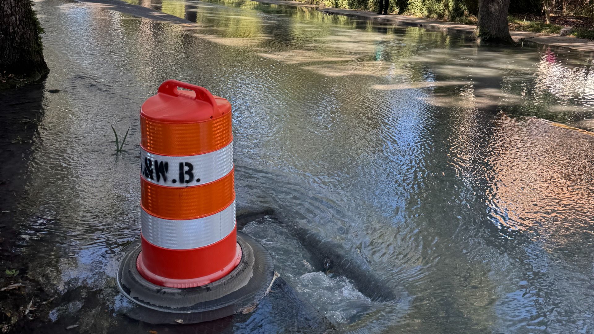 An orange and white construction barrel sits in floodwater on a street, water ripples around its base, with trees and reflections along the edge.