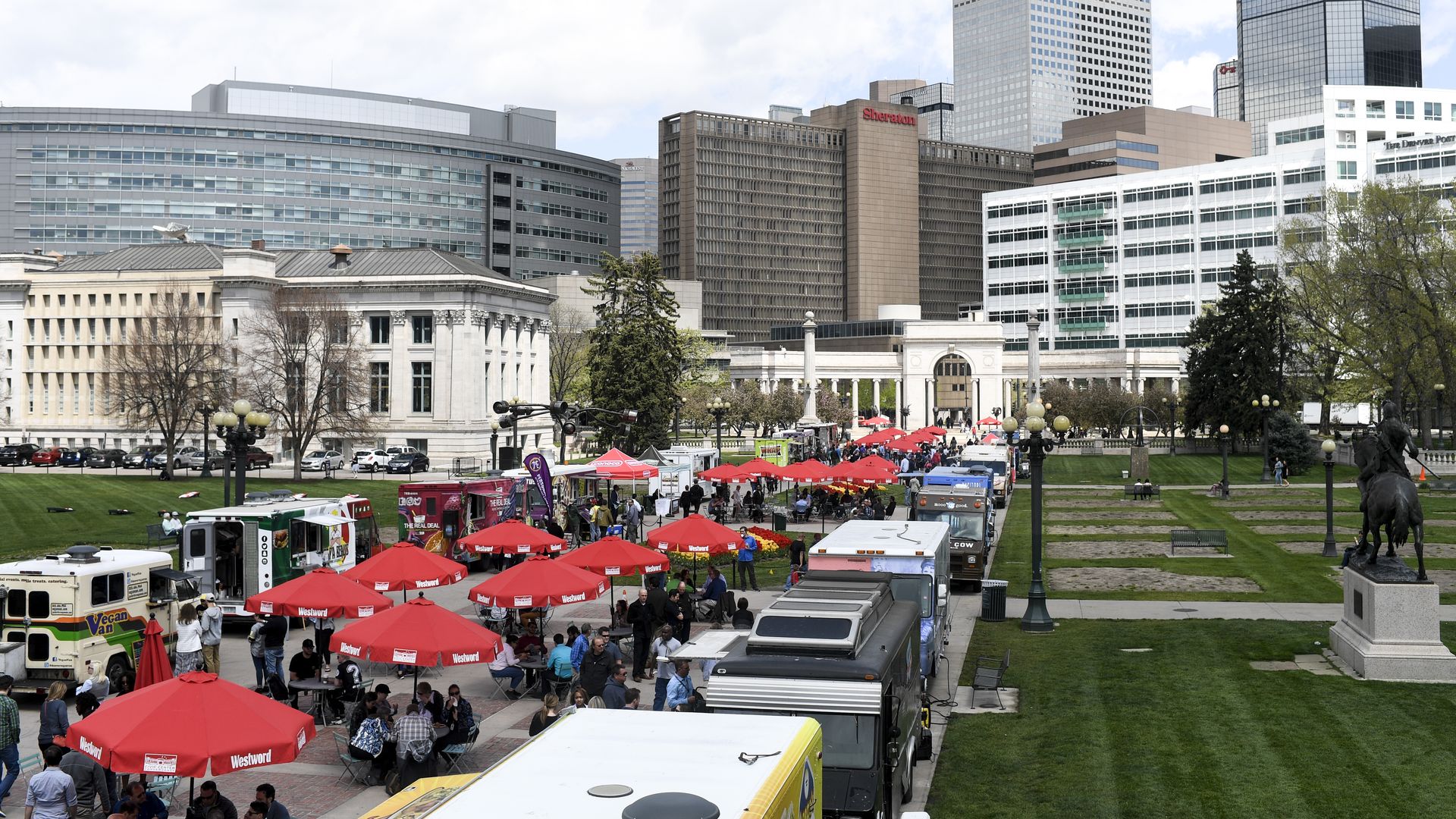 City skyline with tall glass towers, a grassy park in the foreground, and a busy outdoor market with red Westword tents and food trucks; people stroll near a historic building and a statue.