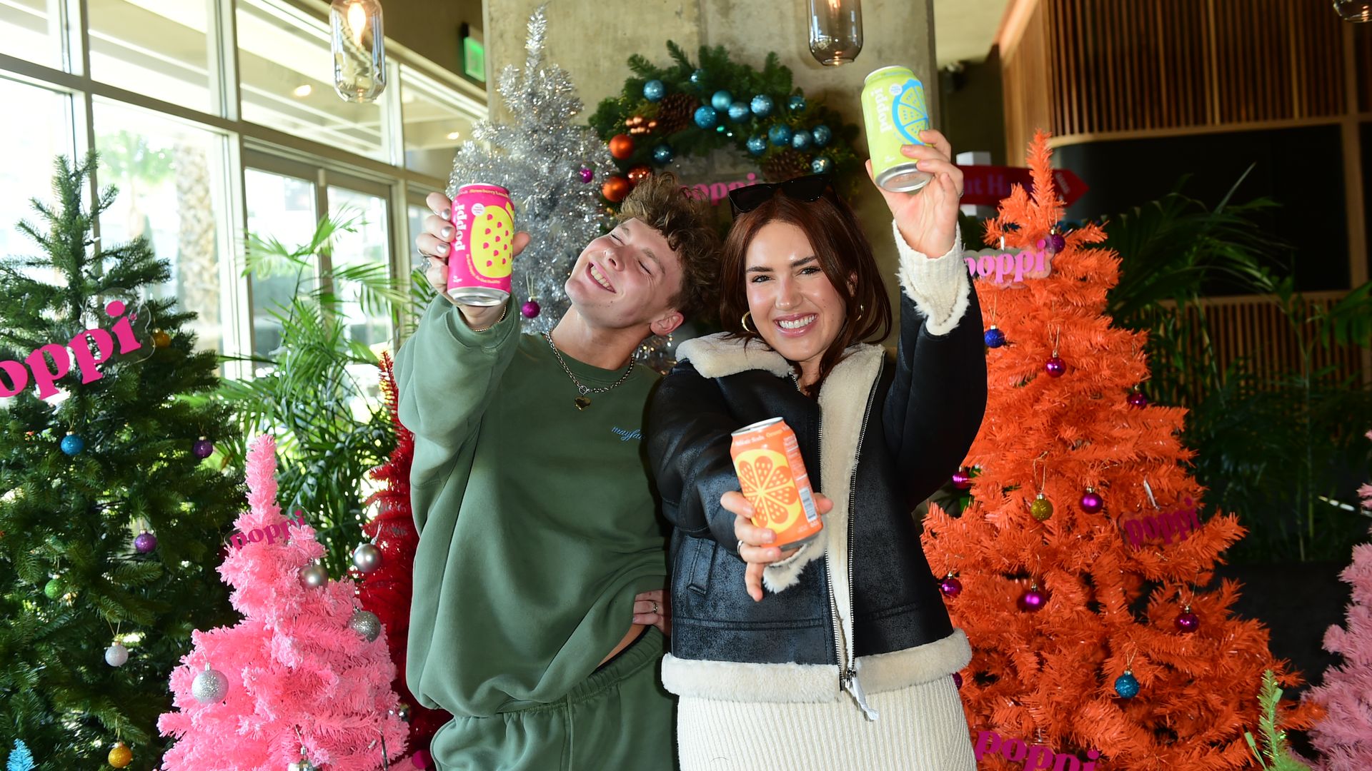 Tik Tok stars Lars Gummer and Tinx, from left to right, appear before Christmas trees holding cans of Poppi-branded soda.