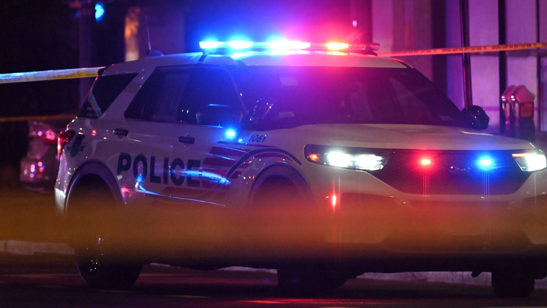 A dc police car with red and blue lights on at night, parked, with a blurry yellow crime scene tape.