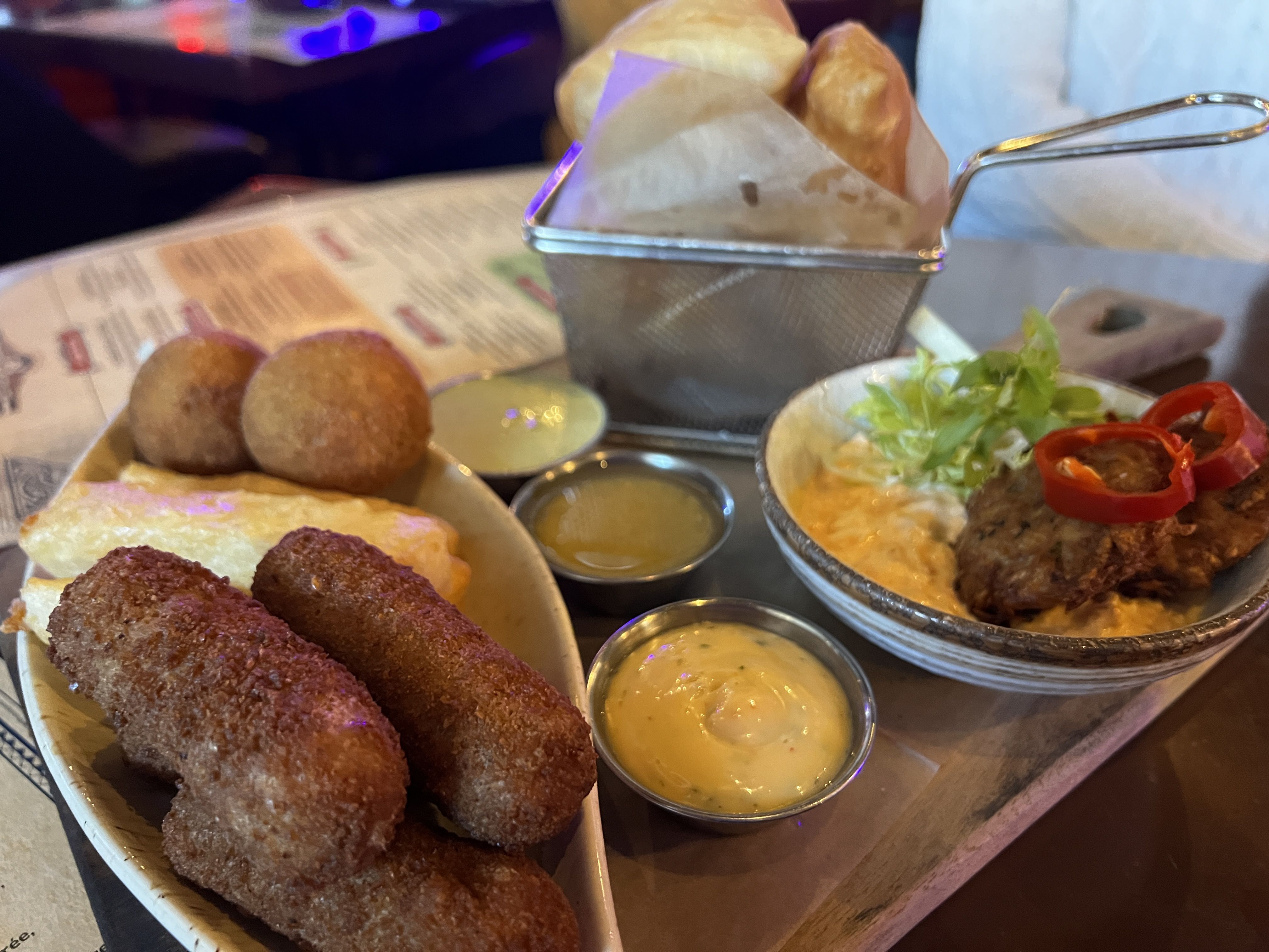 Metal basket with bread and napkin sits on a table; in front, several fried croquettes with yellow dipping sauce cups. A side bowl shows salad greens and red peppers under warm restaurant lighting.