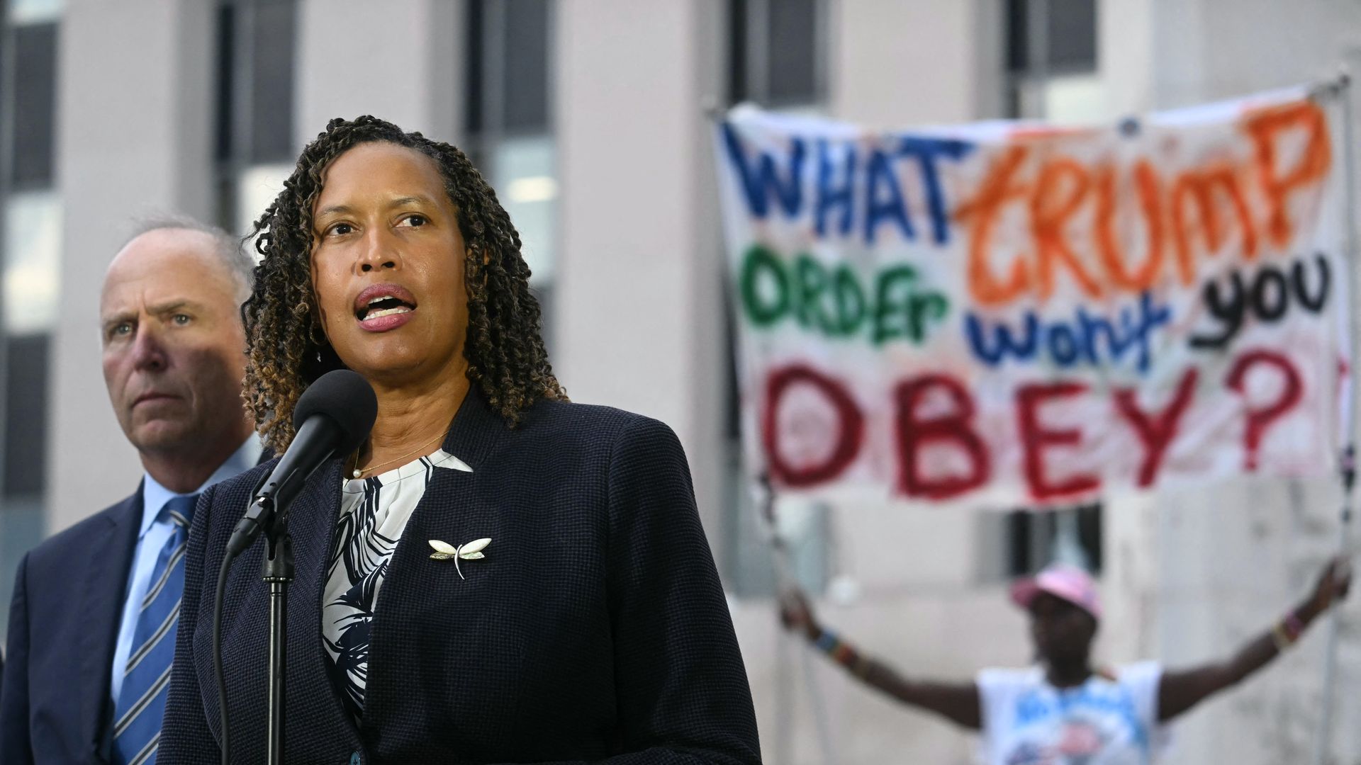 Mayor Bowser -- wearing a black blazer -- speaks at a microphone, a man in a suit stands behind her. In the background, a person holds a colorful sign reading "What Trump order won't you obey?"