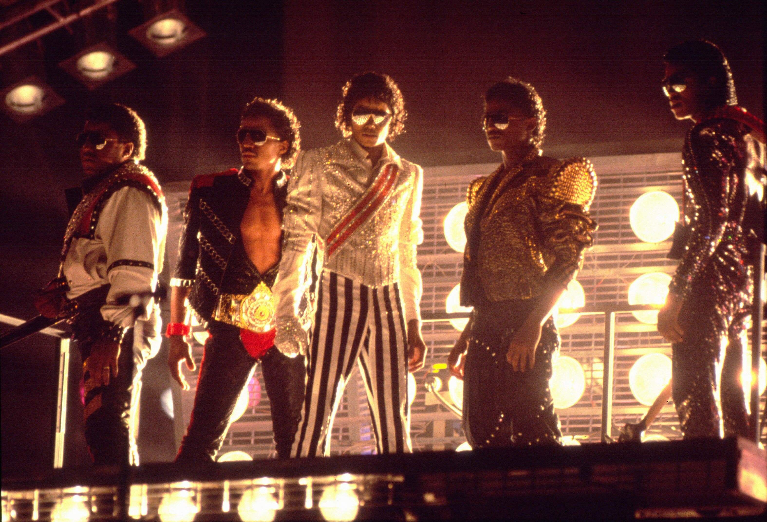 A group of five men in glittering jackets and sunglasses pose on a lit stage; center figure wears a silver sequined shirt with striped pants, others in gold and black, round lights in the background.