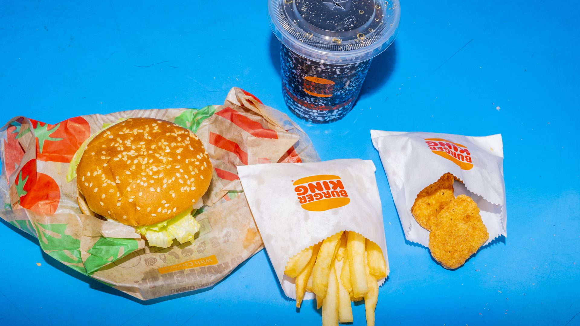 Burger King food on a blue background. Food includes a burger on top of a wrapper, pack of fries, nuggets and soda in clear plastic cup