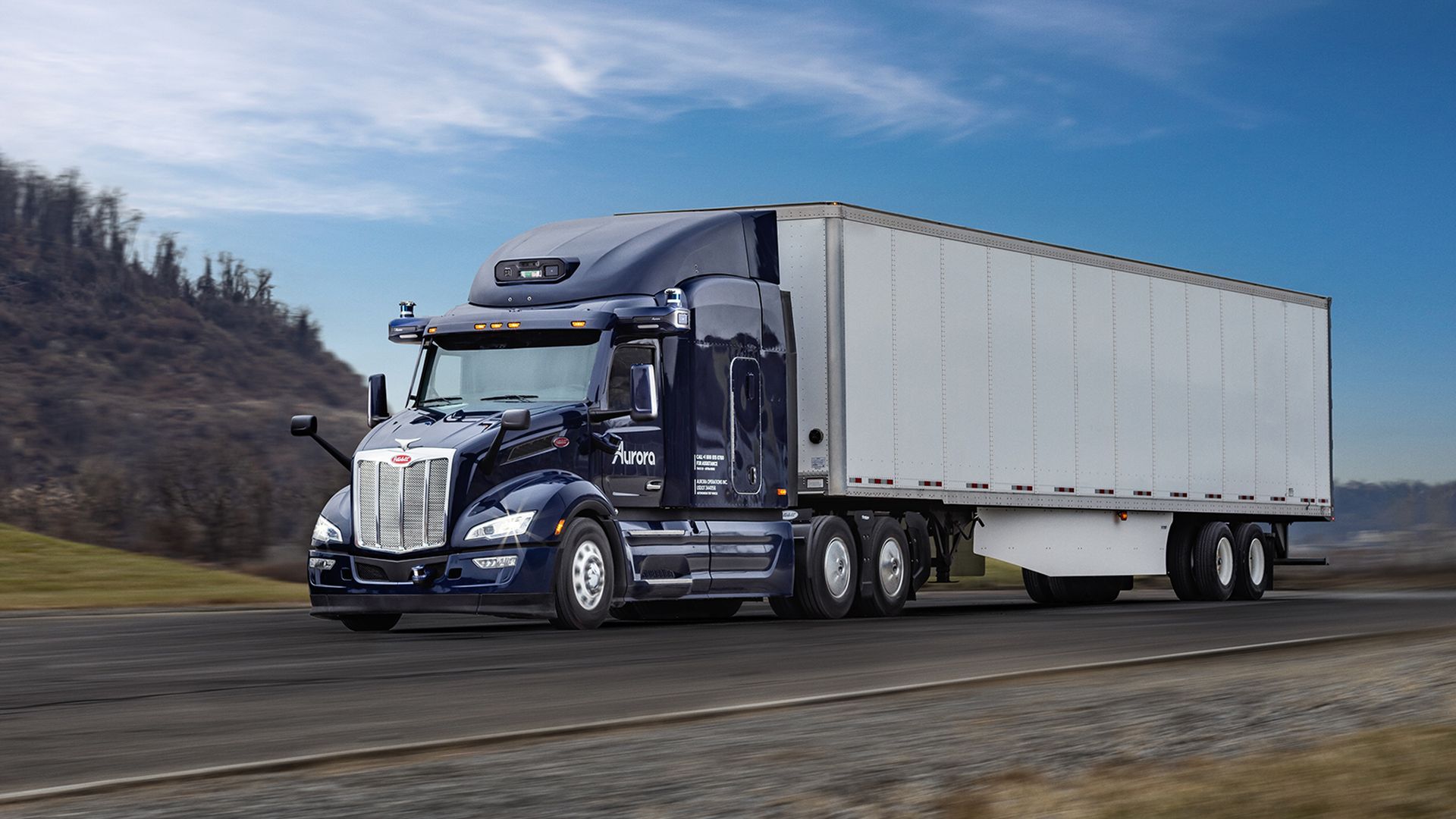 A dark blue semi-truck with a long silver trailer speeds along a highway, with motion blur on the road and distant hills under a clear blue sky. Chrome grille, side mirrors, and amber lights are visible.