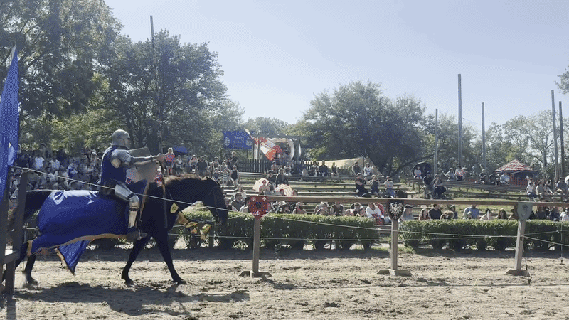 A knight in blue armor rides a blue-draped horse through a fenced arena at a medieval reenactment, with spectators on wooden bleachers watching under a bright sunny sky.