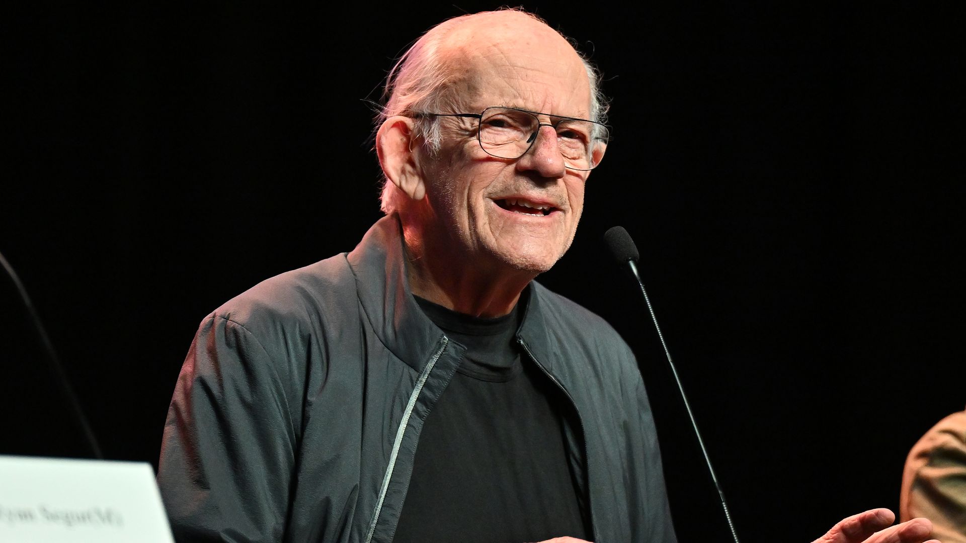 Actor Christopher Lloyd speaks into a microphone on a stage with a black backdrop. 