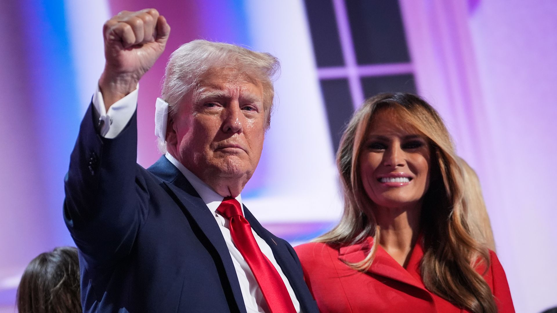 Donald Trump stands on the left of the photo with one ear covered by a bandage and a hand in the air. Melania Trump stands next to him, to the right of the photo, smiling. 