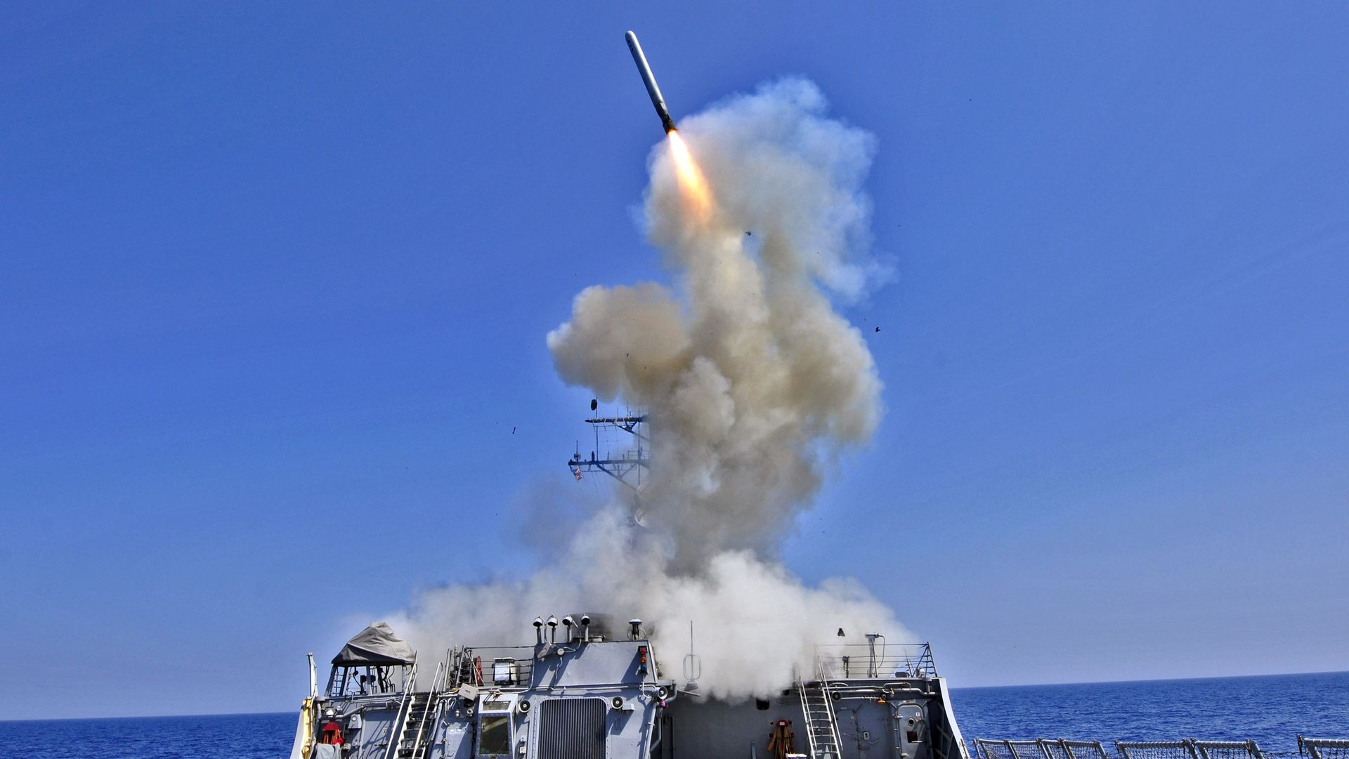 A missile launches upward from a gray naval ship deck, surrounded by thick white smoke under a clear blue sky over the ocean.