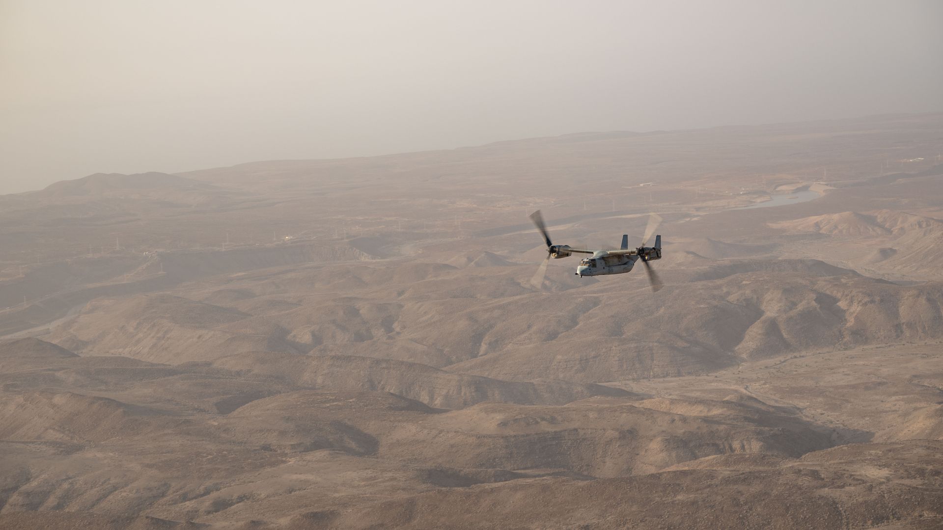 A gray military tiltrotor aircraft, the MV-22 Osprey, flies low over a hazy desert basin with rolling brown hills and a winding river in the distance.
