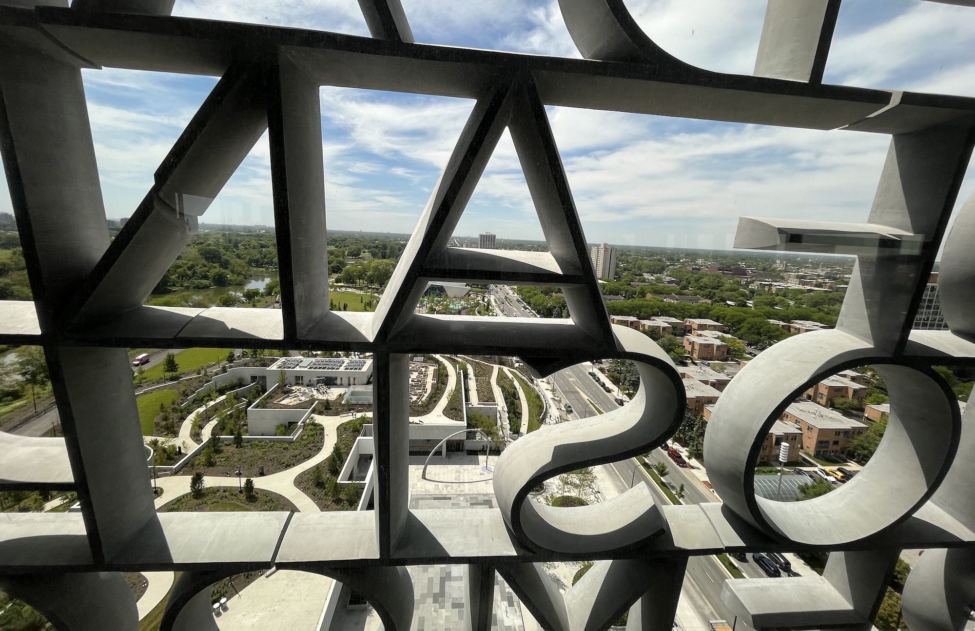 View through a bold geometric metal lattice with square and triangular openings, revealing a cityscape: green park, winding roads, river, and distant buildings under a blue sky with clouds.